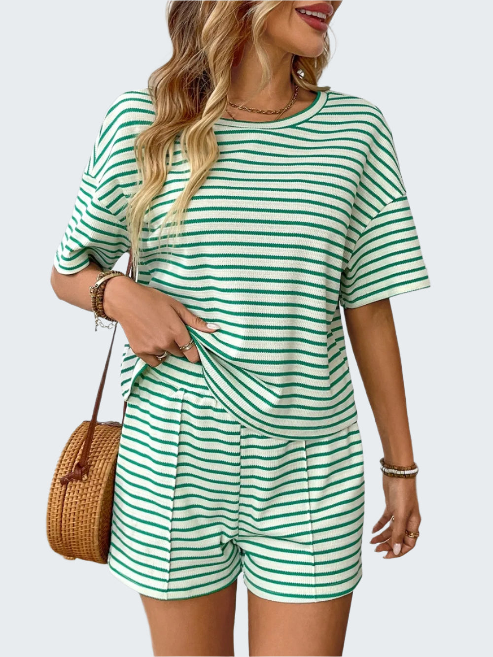 A woman wearing a green and white striped round neck top with short sleeves and matching shorts, accessorized with a straw bag and a watch.