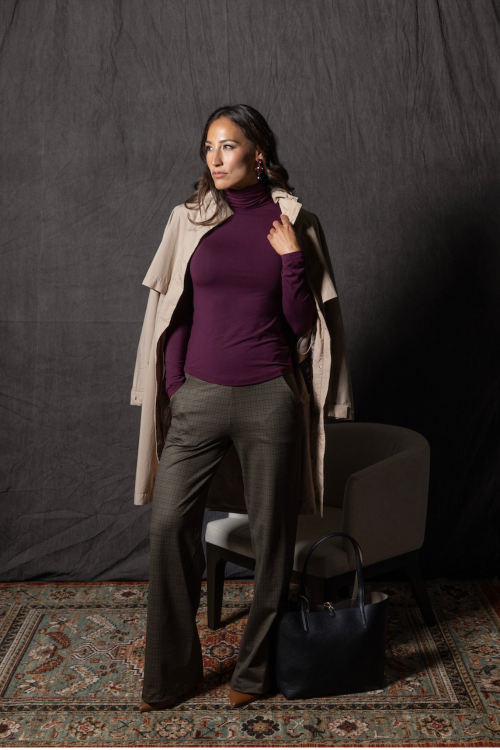 Woman in a purple turtleneck and beige coat standing against a dark background