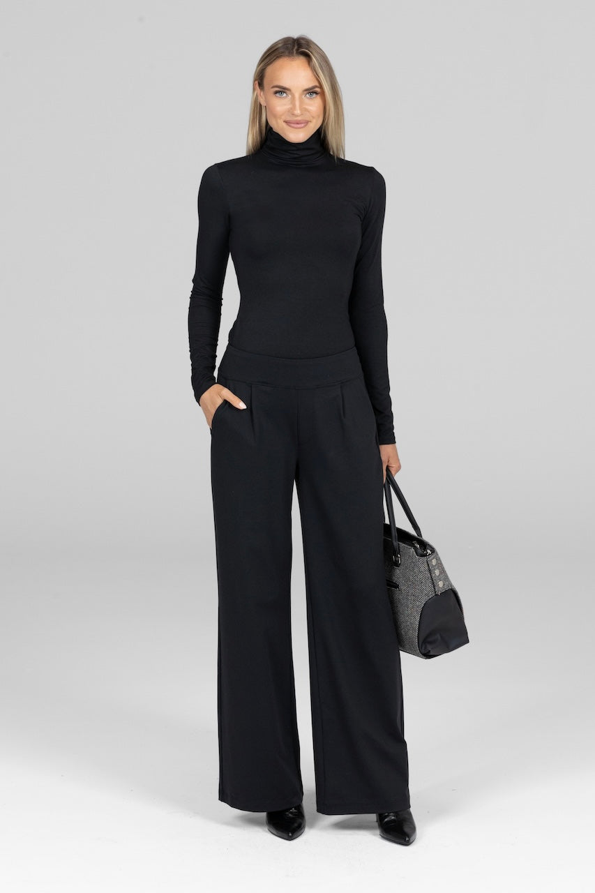 Woman wearing a black turtleneck and wide-leg pants holding a black handbag on a white background