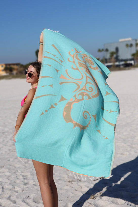 Woman holding a turquoise beach towel with gold patterns on a sandy beach.