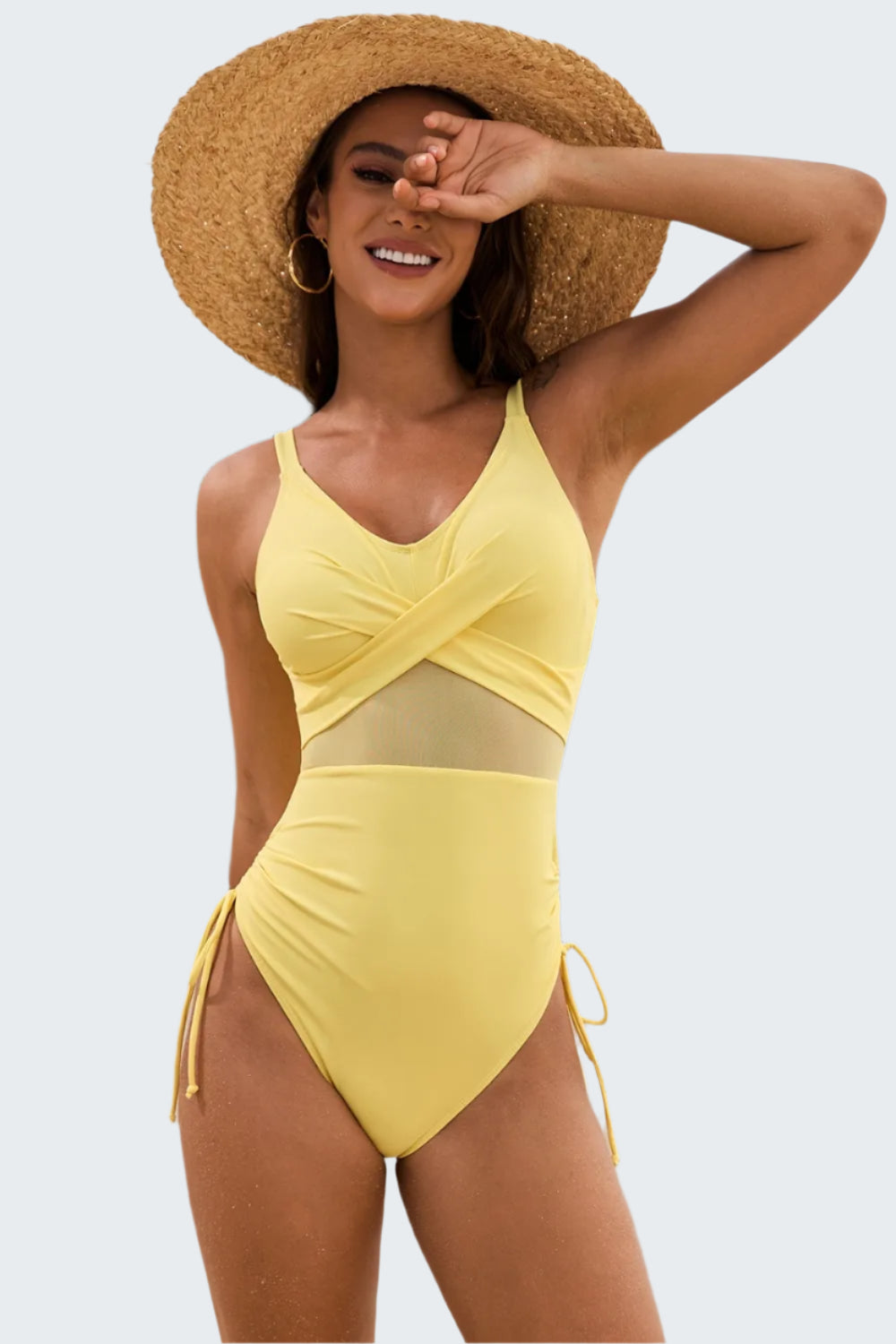 A woman at the beach wearing a yellow one-piece swimsuit with a v-neck and drawstrings. She is also wearing a wide-brimmed straw hat.