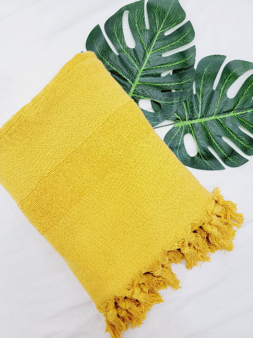 Yellow towel with tassels on a white background with a monstera leaf.