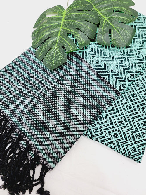 Textile with green and black striped pattern and tassels, placed on a white surface with green leaves.