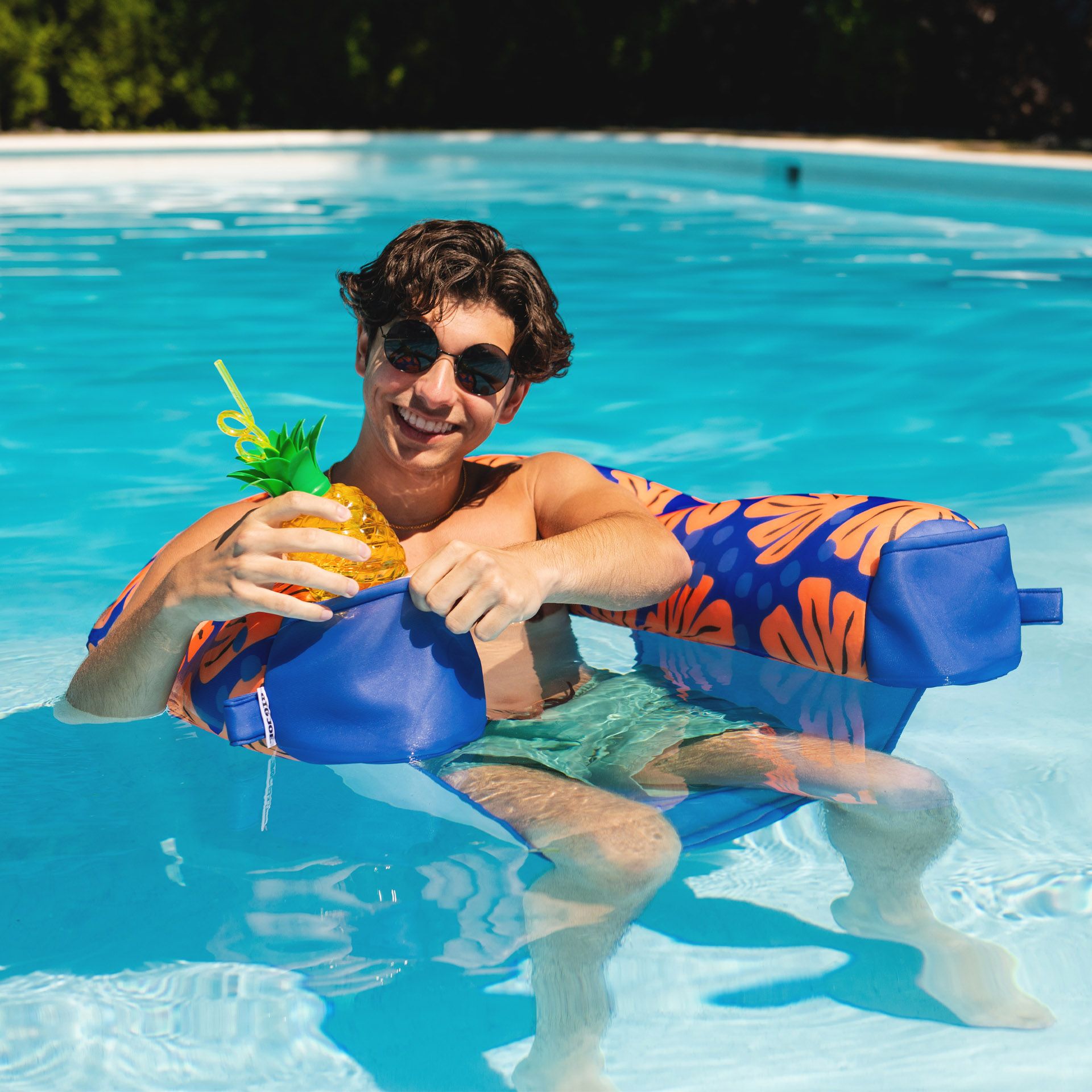 Person sitting on a colorful inflatable pool float holding a pineapple drink in a pool.
