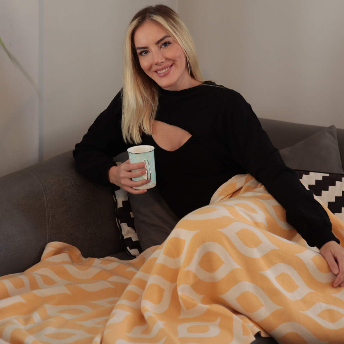 Woman sitting on a couch with a yellow patterned blanket and holding a cup.