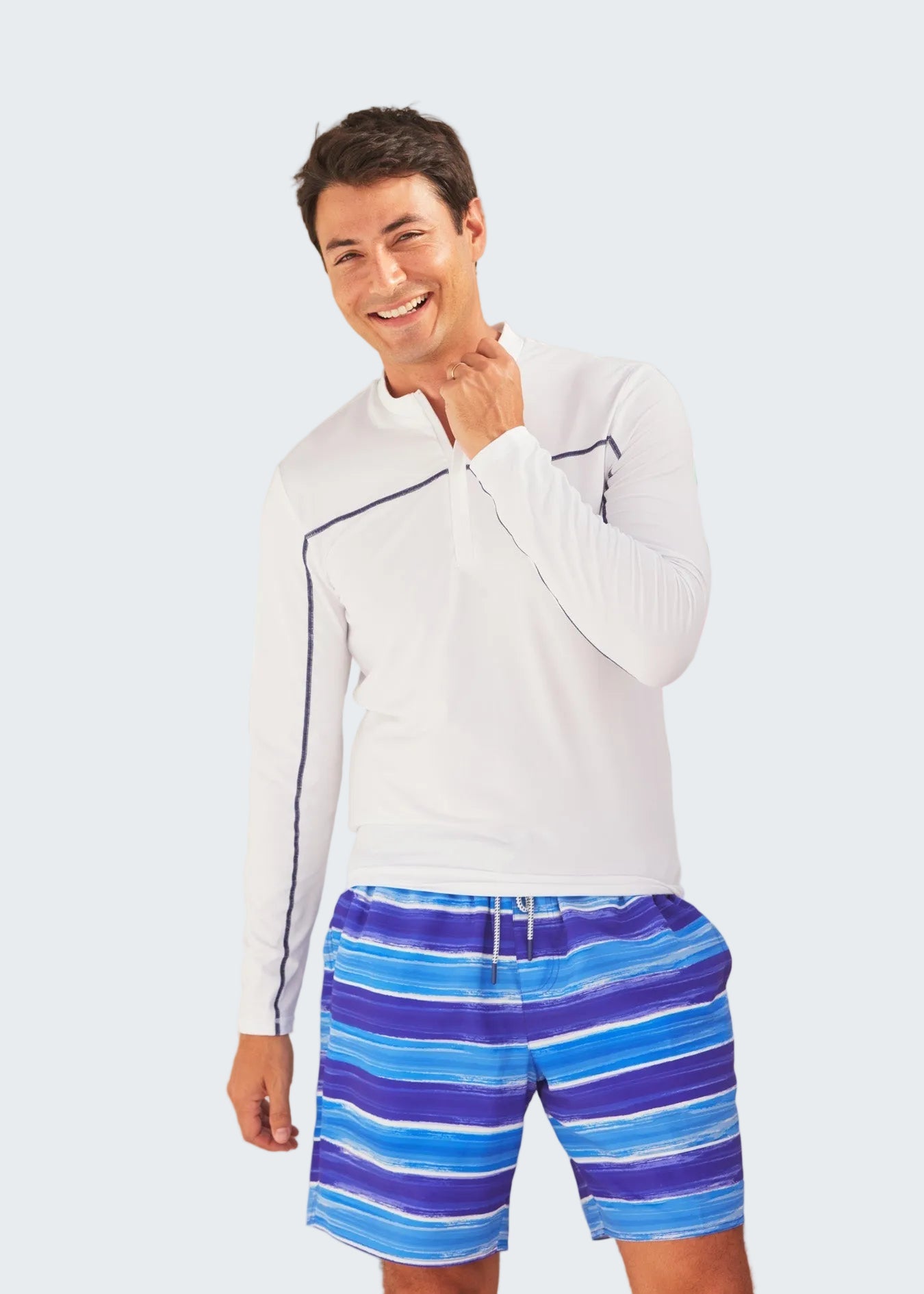 Man wearing a white long-sleeve shirt and blue striped shorts standing in front of a polka dot surfboard.