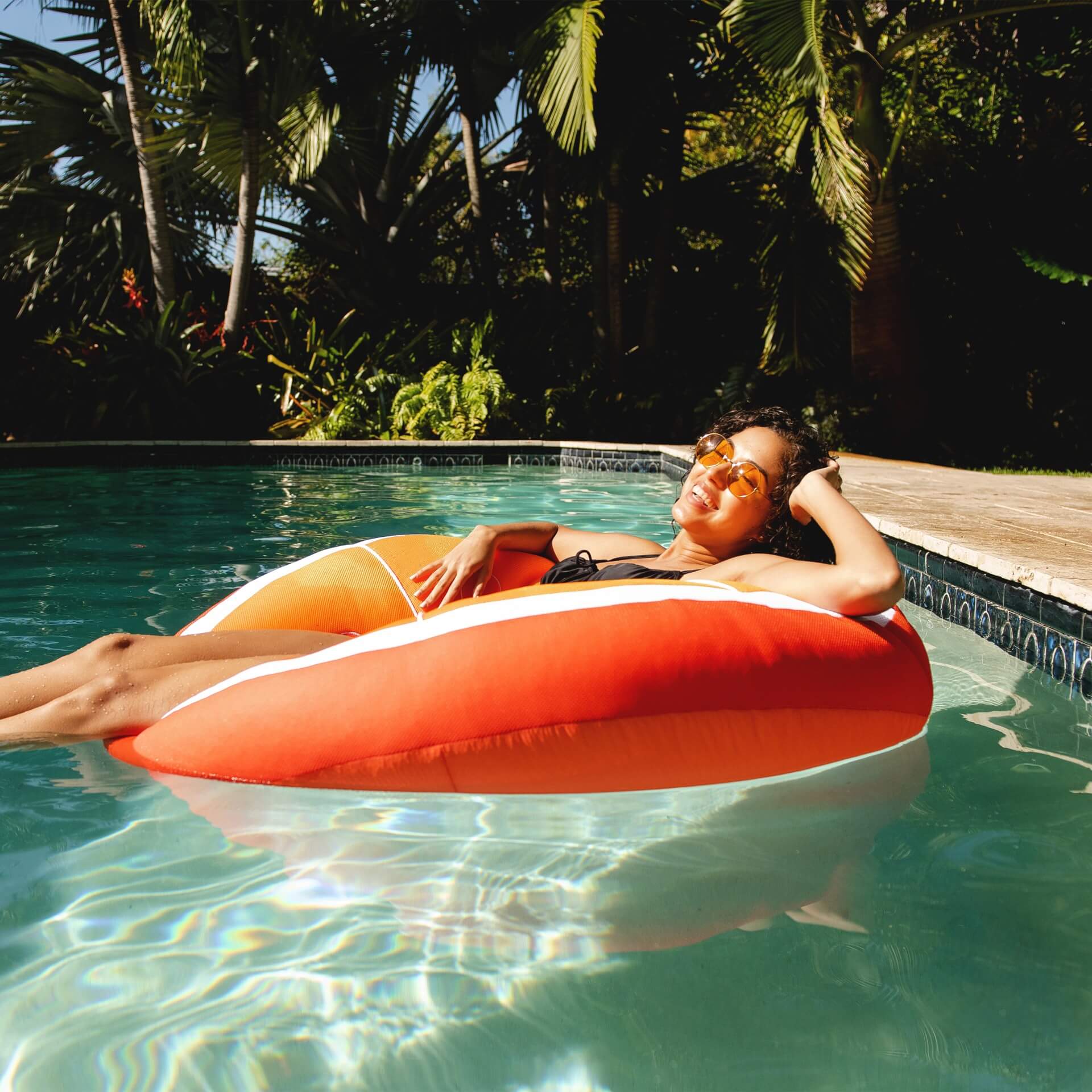 Person lounging on an orange inflatable ring in a pool surrounded by lush greenery