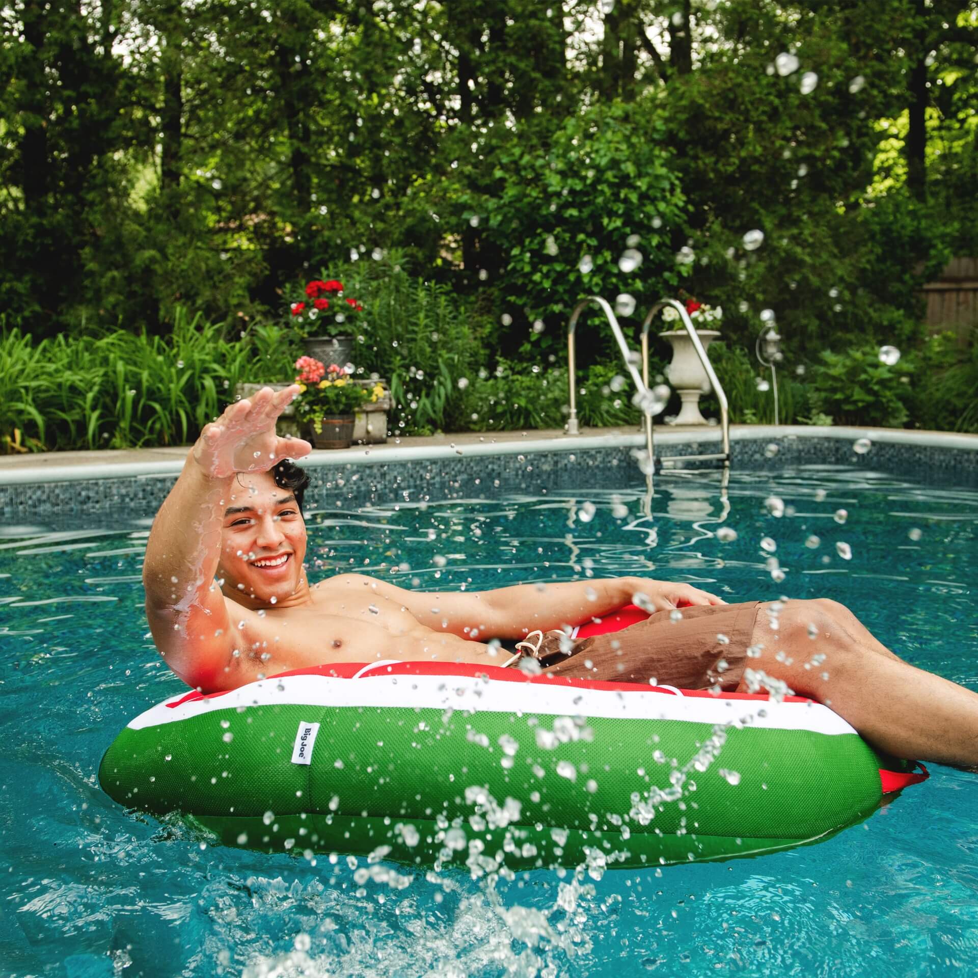 Person lounging on a green and red inflatable raft in a pool with trees and flowers in the background