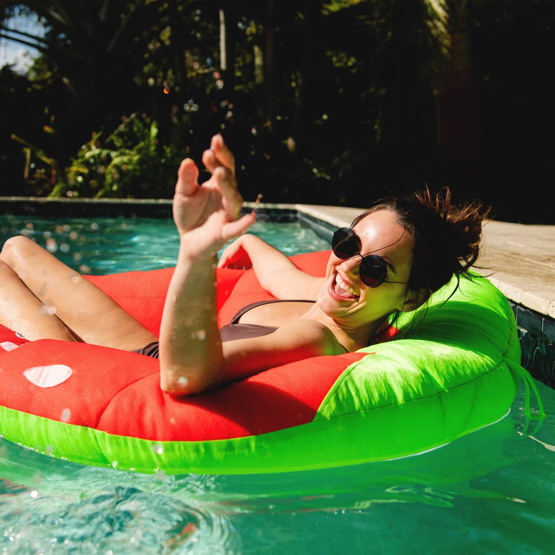 Woman lounging on a watermelon-shaped inflatable pool float in a pool with trees in the background