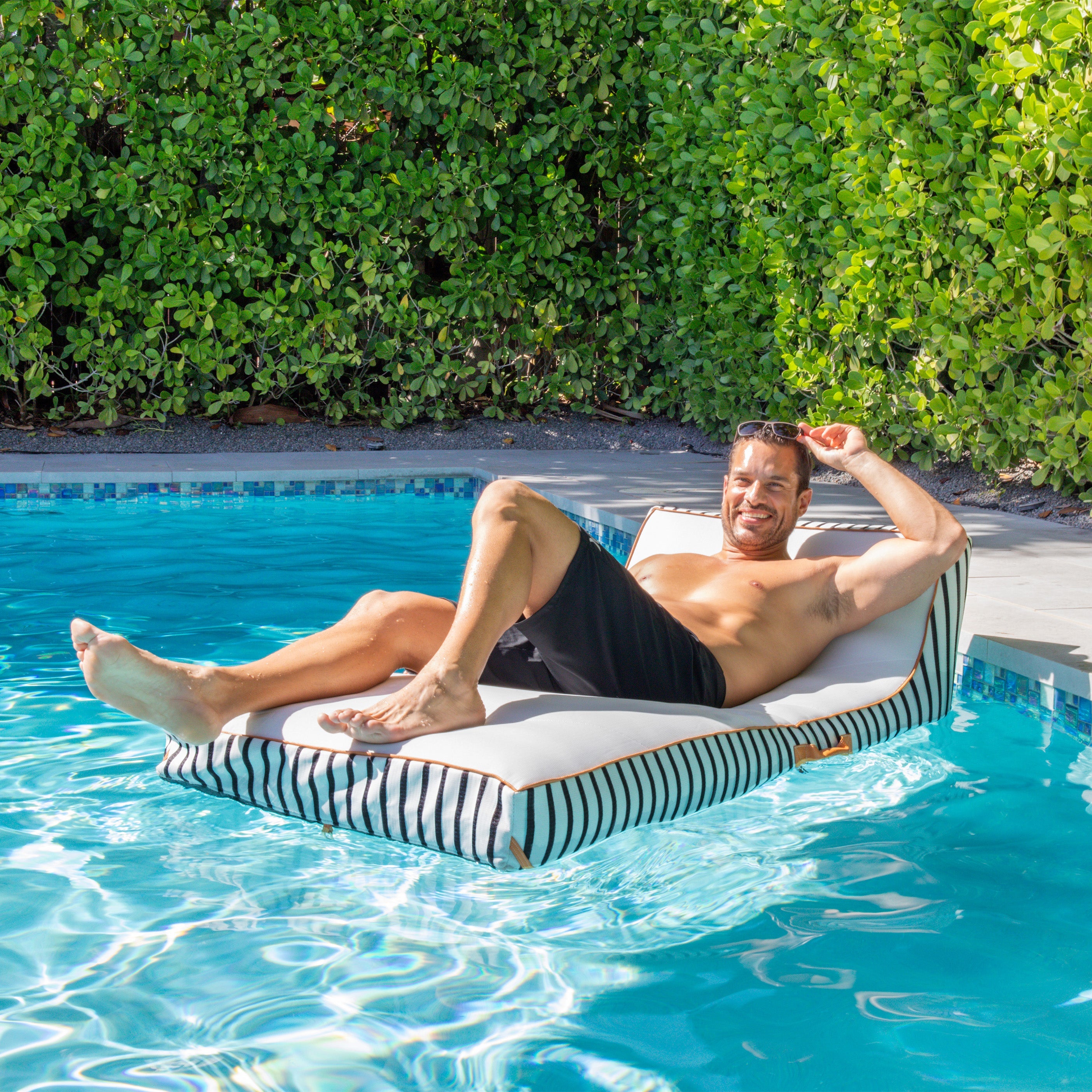 Man relaxing on a striped pool float in a swimming pool with greenery in the background