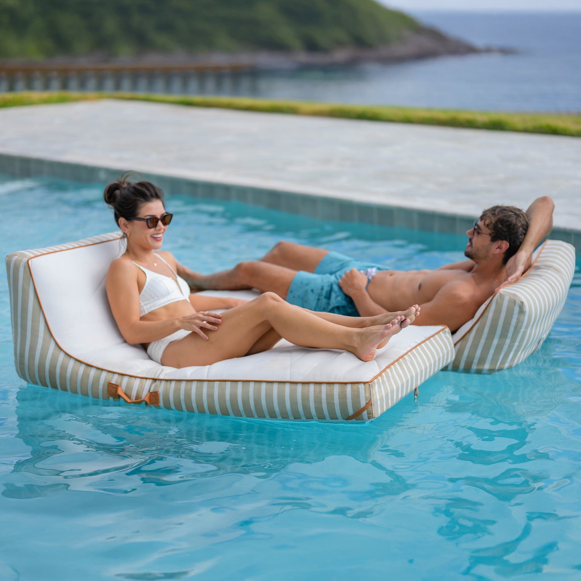 Two people relaxing on a striped inflatable pool float in a pool with a scenic background.