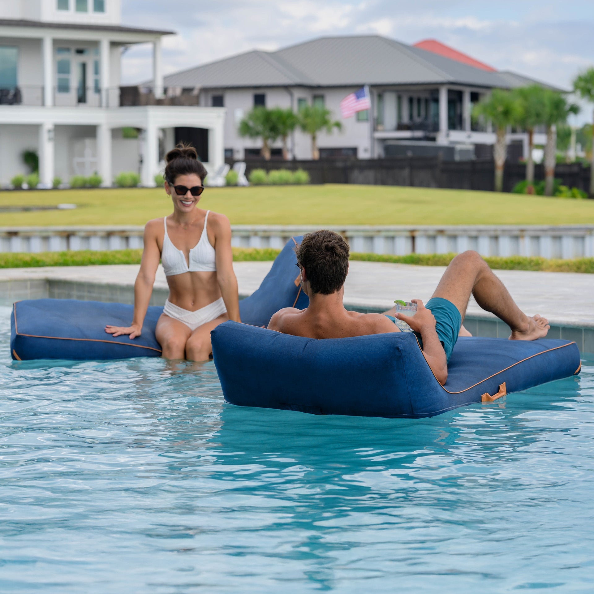 Two people lounging on inflatable blue lounge chairs in a pool with houses and greenery in the background.