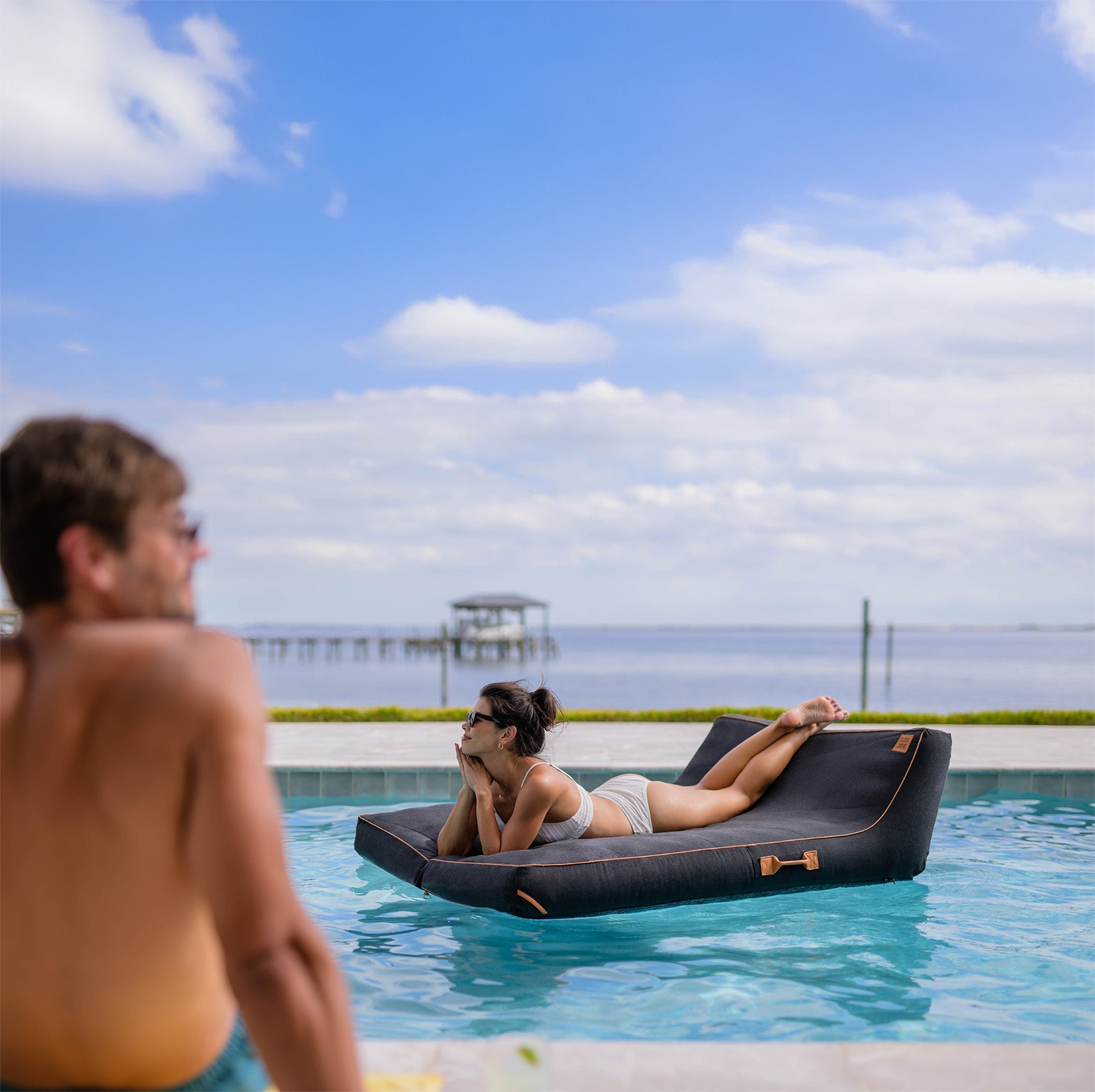 Woman lying on a pool float in a pool with a man watching her, under a clear blue sky.