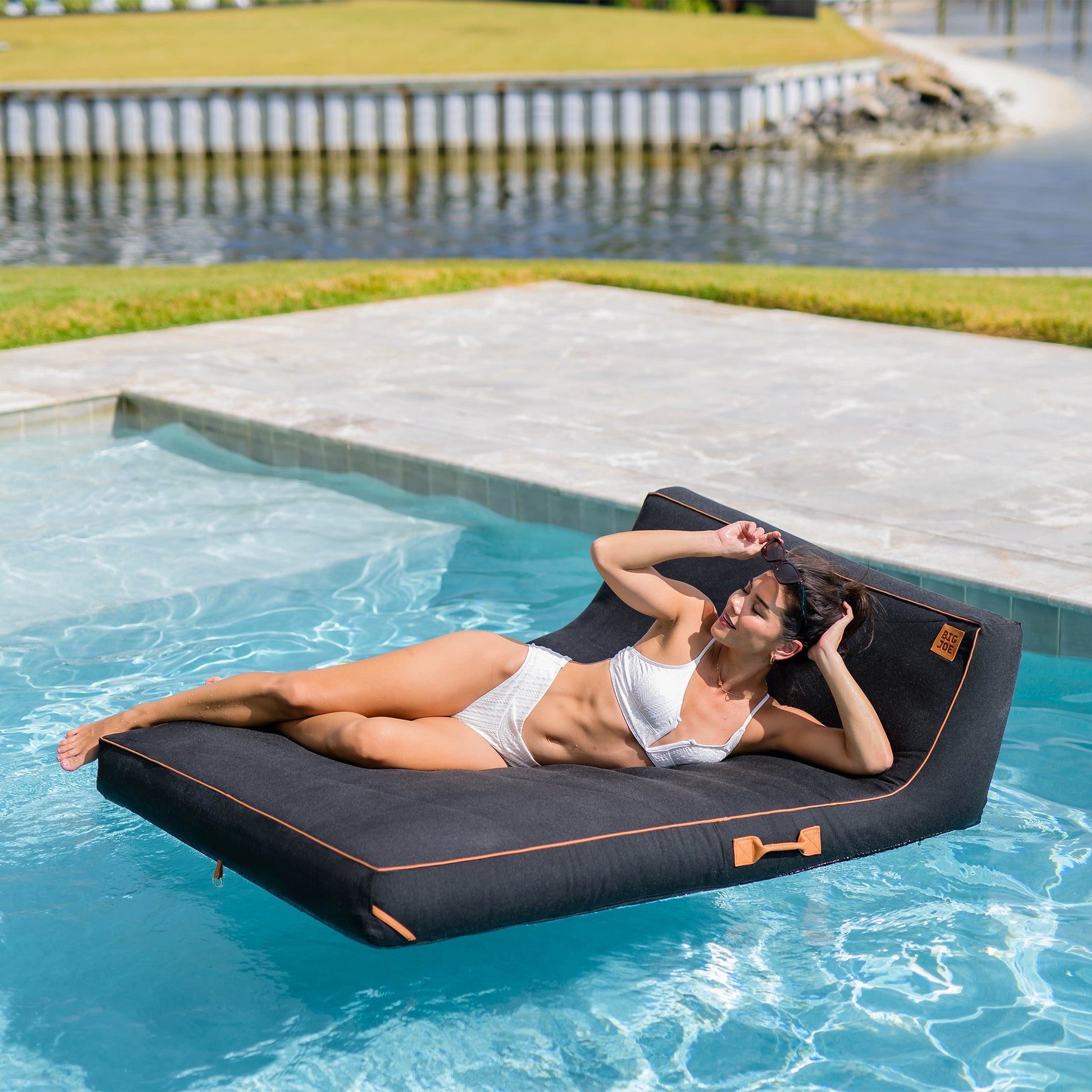 Woman lying on a floating pool mat by a poolside with grass and water in the background