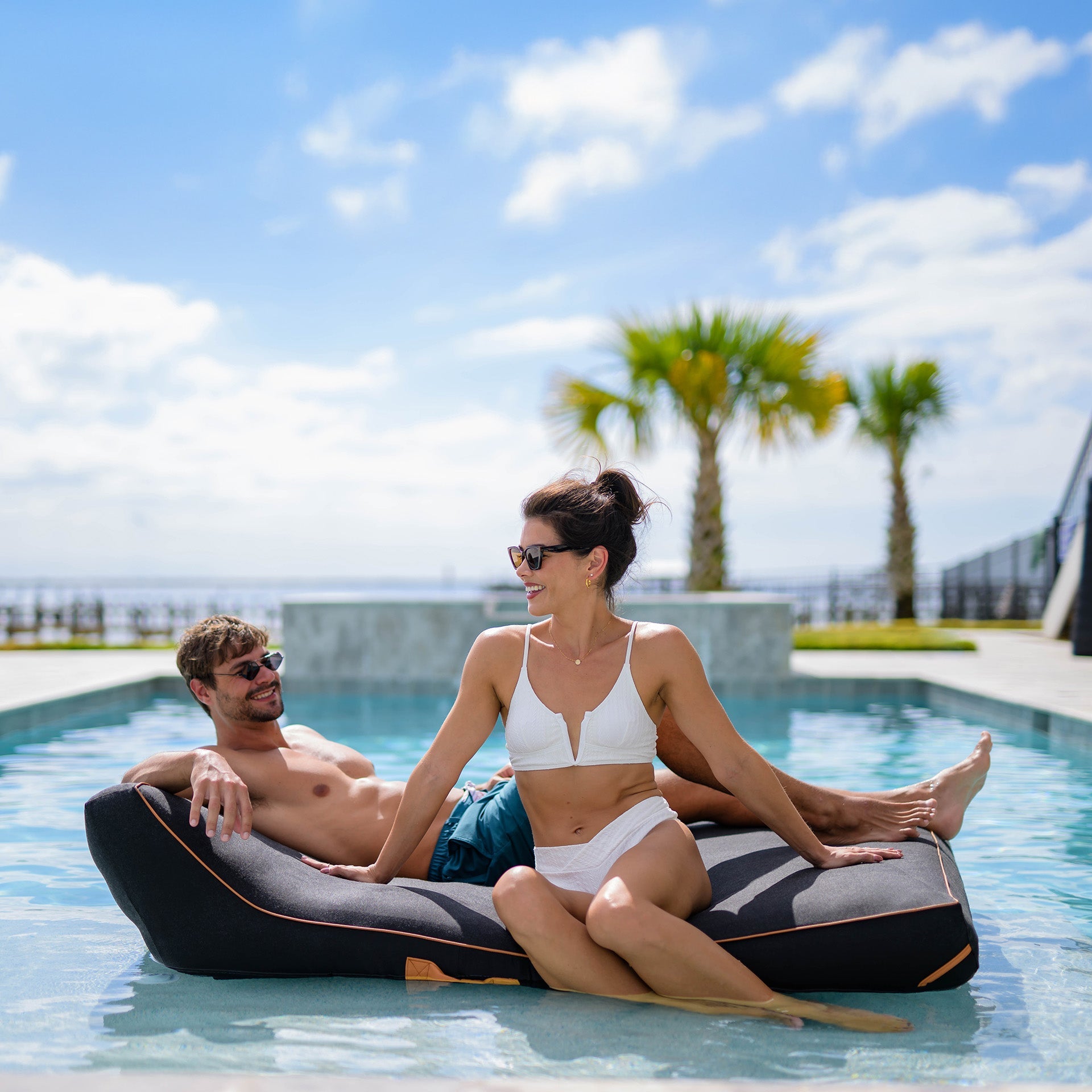 Man and woman relaxing on a pool float in a pool with palm trees in the background