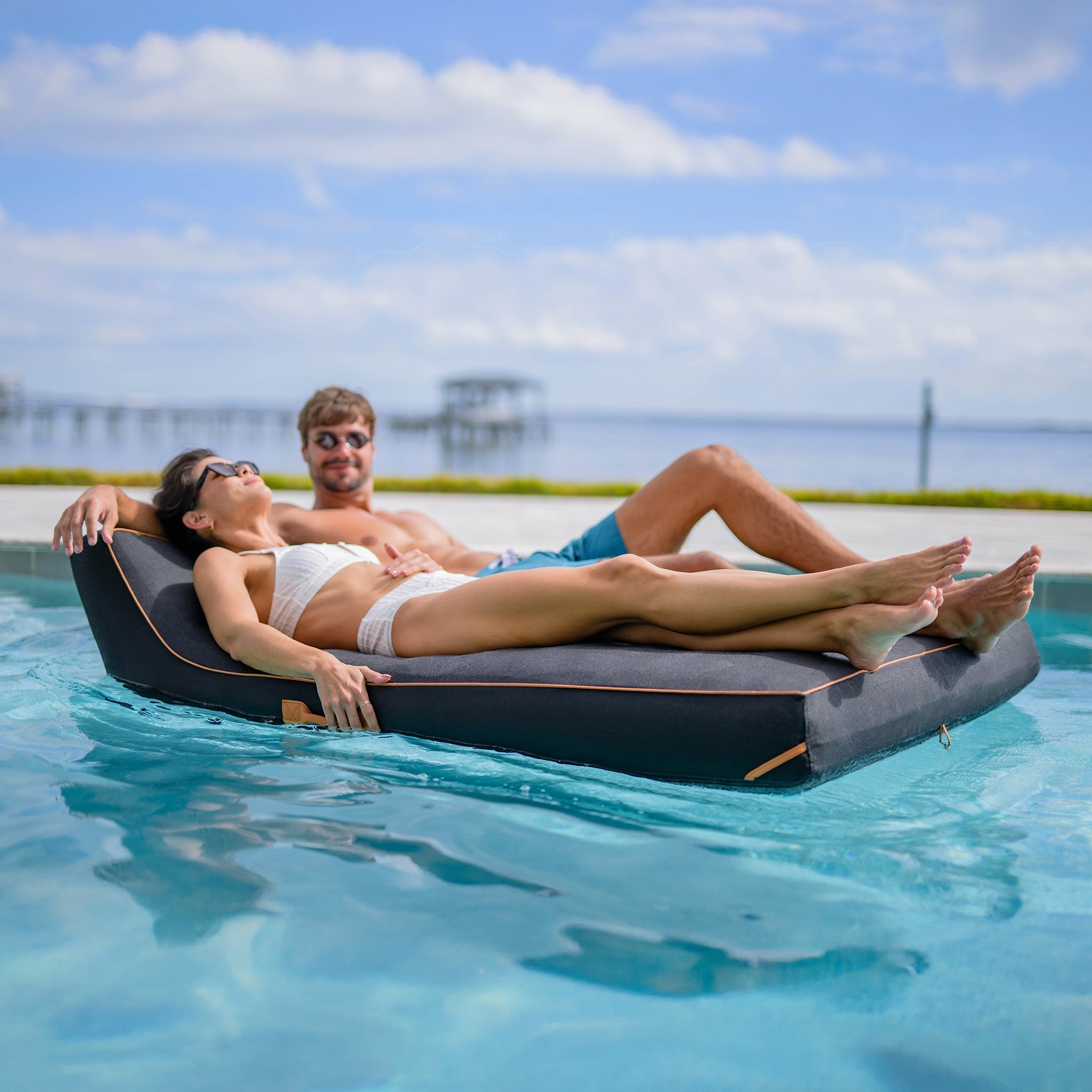 Two people relaxing on a floating platform in a pool with a scenic background.