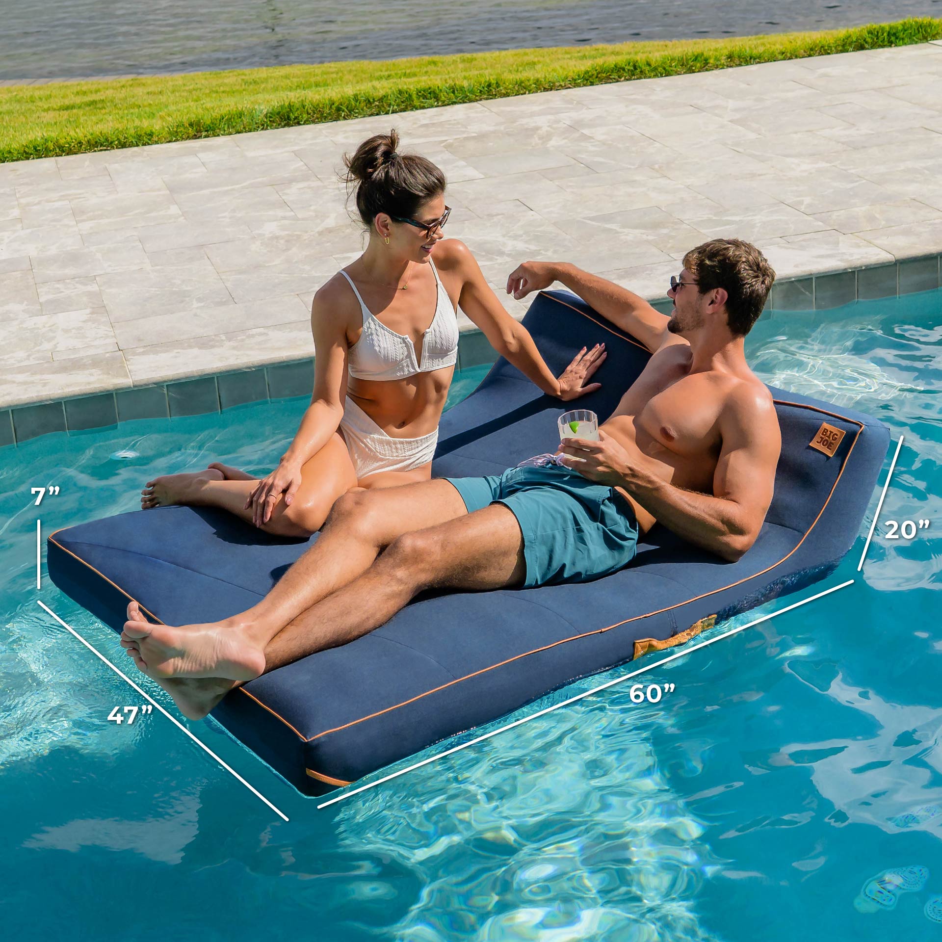Two people relaxing on a blue inflatable pool float with dimensions displayed, by a pool.