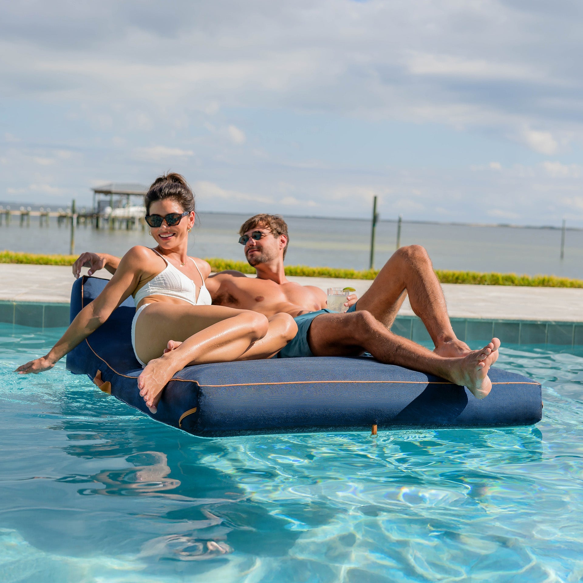 Two people relaxing on a blue inflatable pool float in a pool with a scenic background.