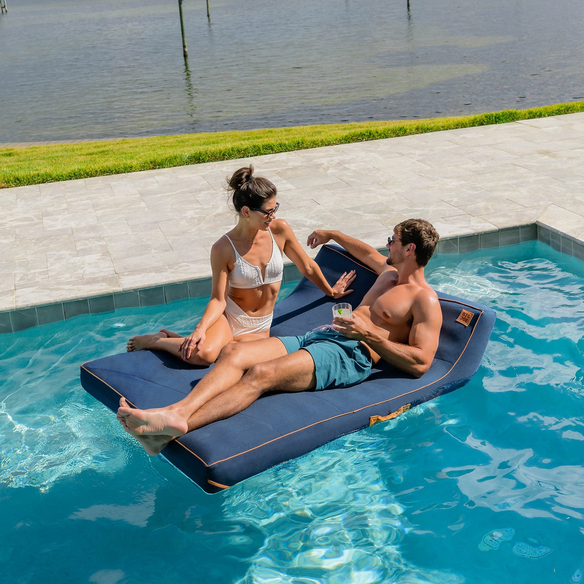 Two people relaxing on a floating mat in a pool with a scenic background