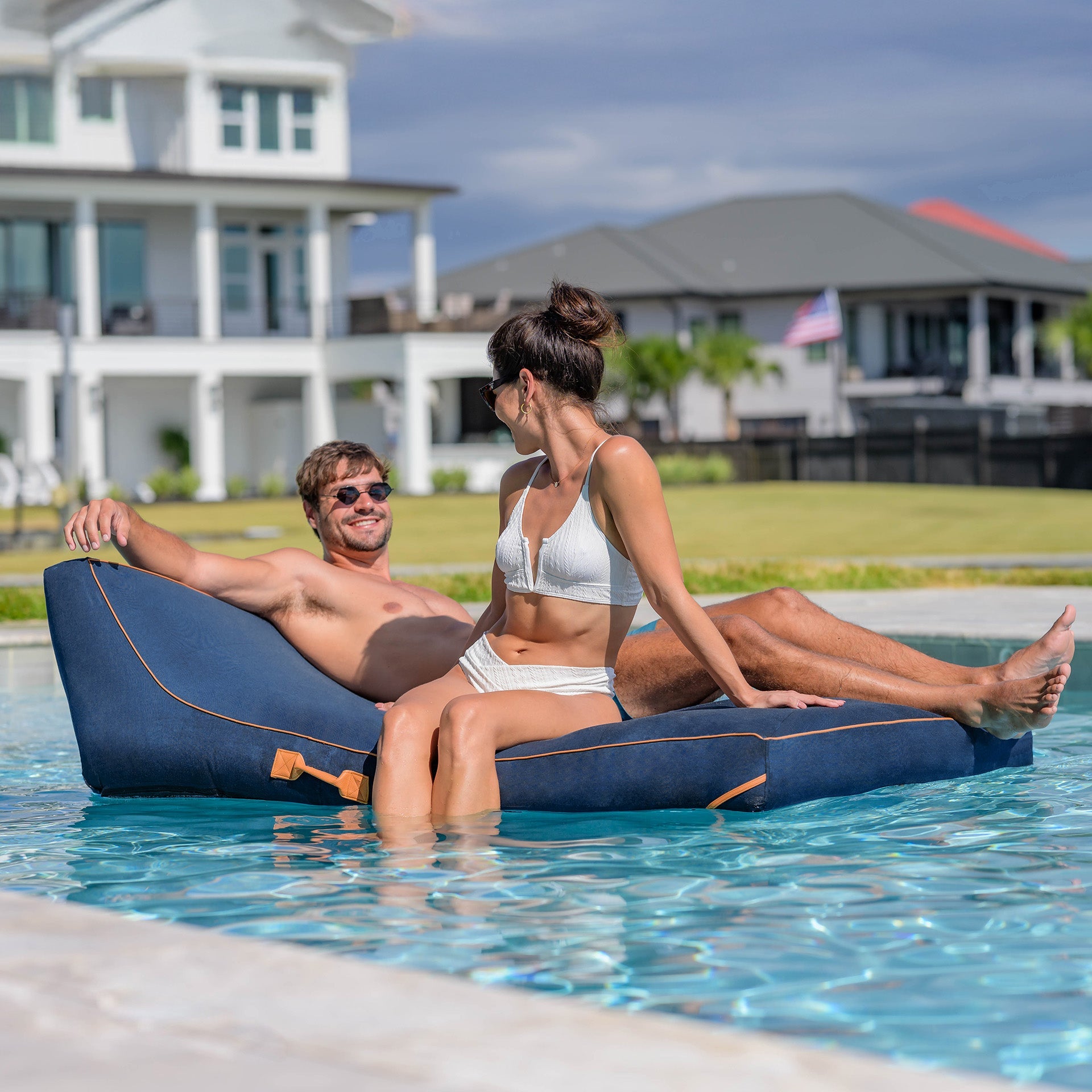 Two people relaxing on a blue inflatable lounger in a pool with a house in the background.