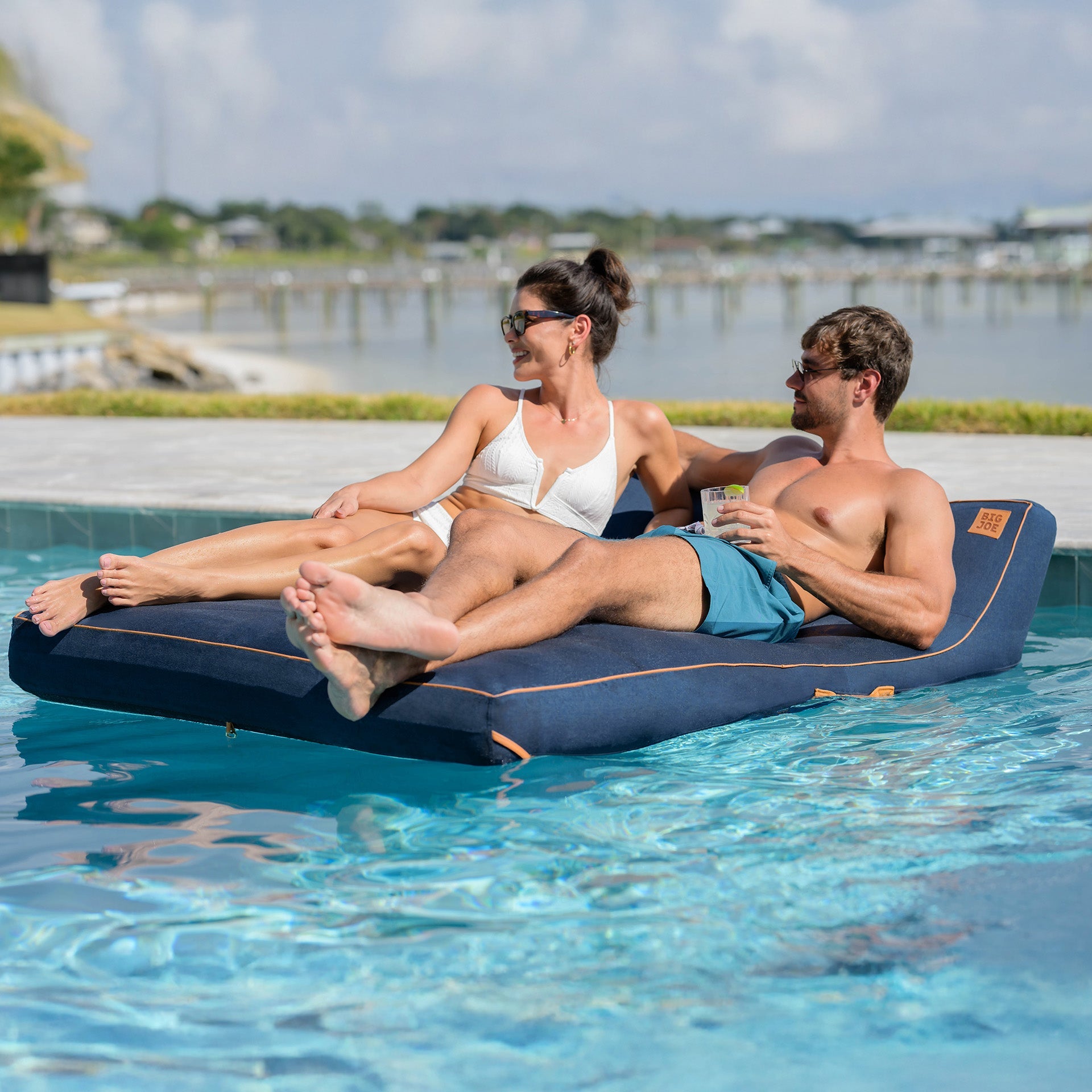 Two people relaxing on a floating device in a pool with a scenic background.