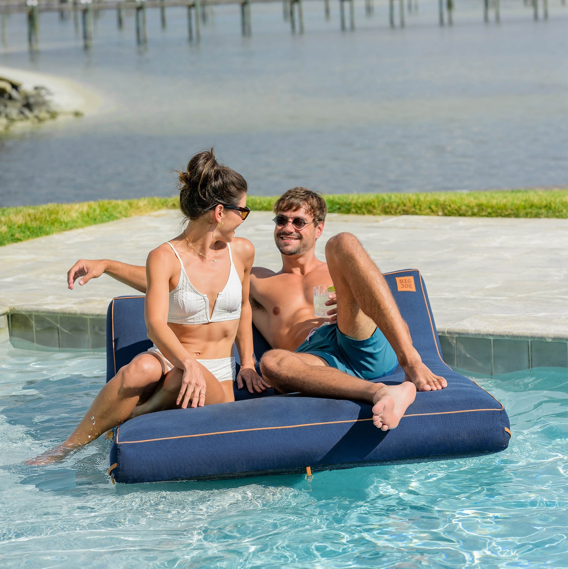 Two people sitting on a blue inflatable pool float by a pool with a scenic background.