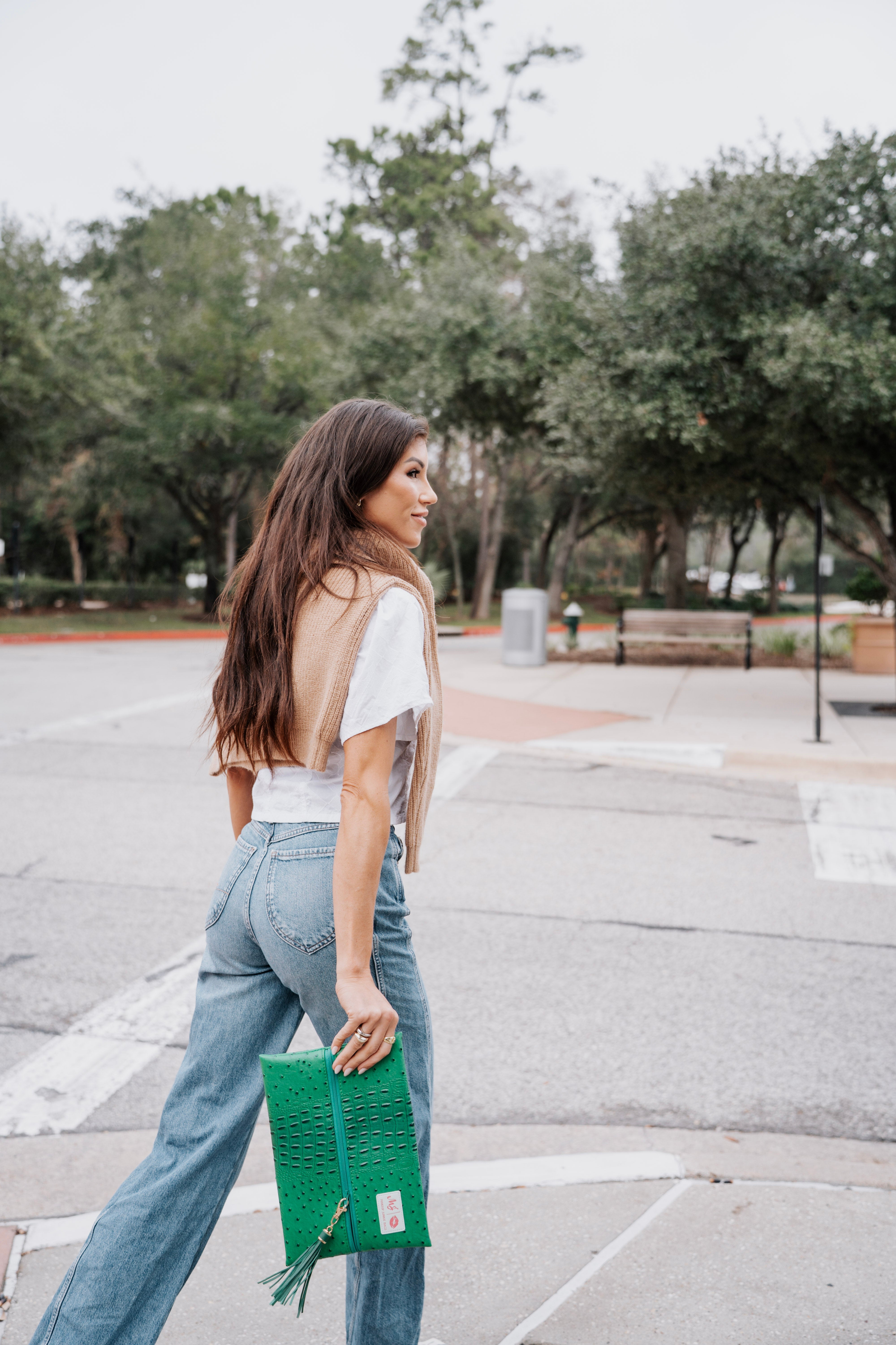 Woman holding a green clutch in an outdoor setting with trees and a bench.