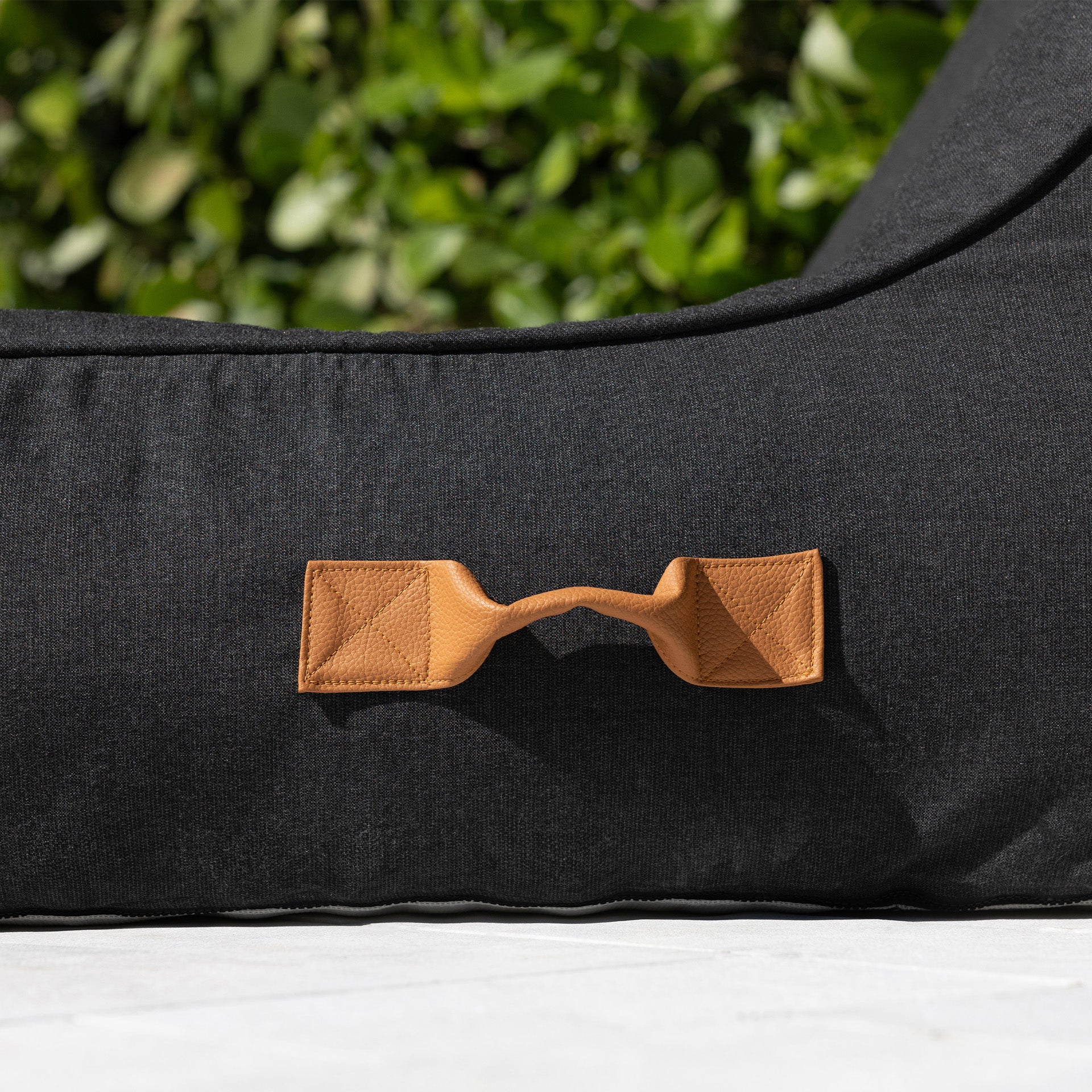 Black dog bed with a brown leather handle against a green outdoor background