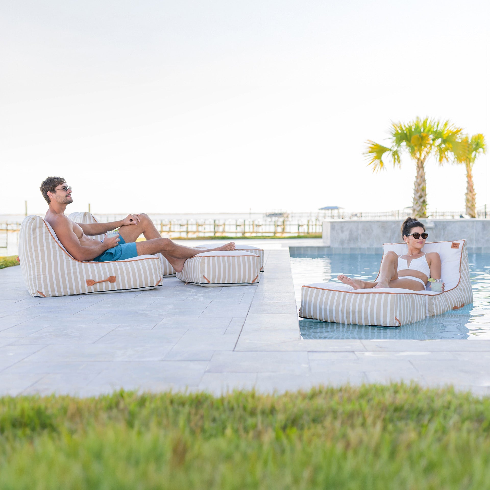 Two people relaxing on striped bean bags by a pool with palm trees in the background.