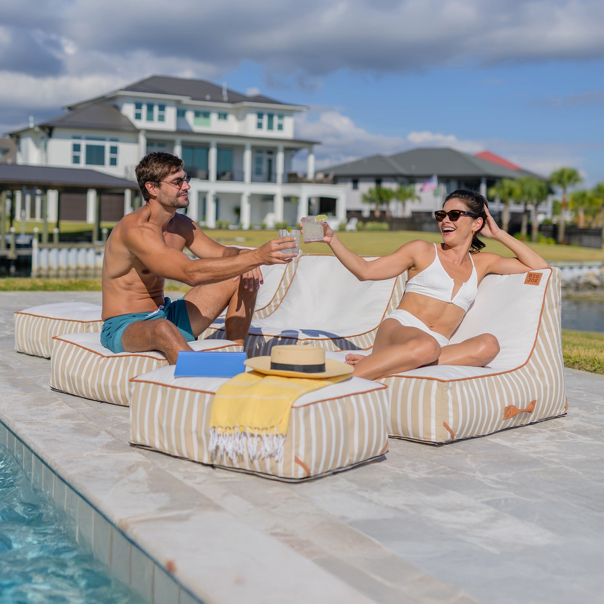Two people sitting on lounge chairs by a pool with a house and palm trees in the background.