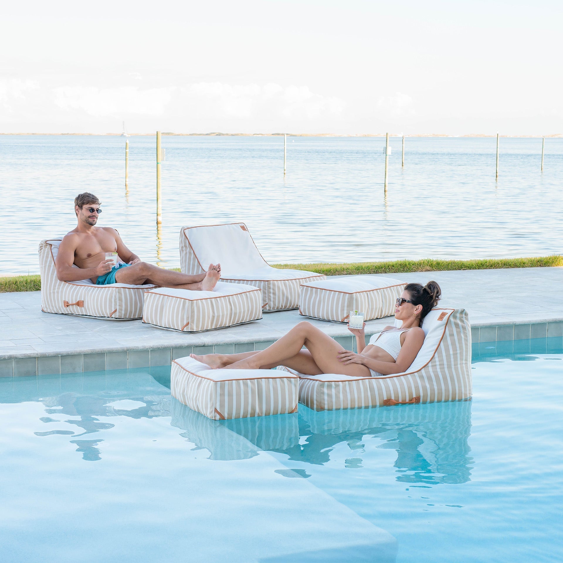 Two people relaxing on striped lounge chairs by a pool with a scenic background.