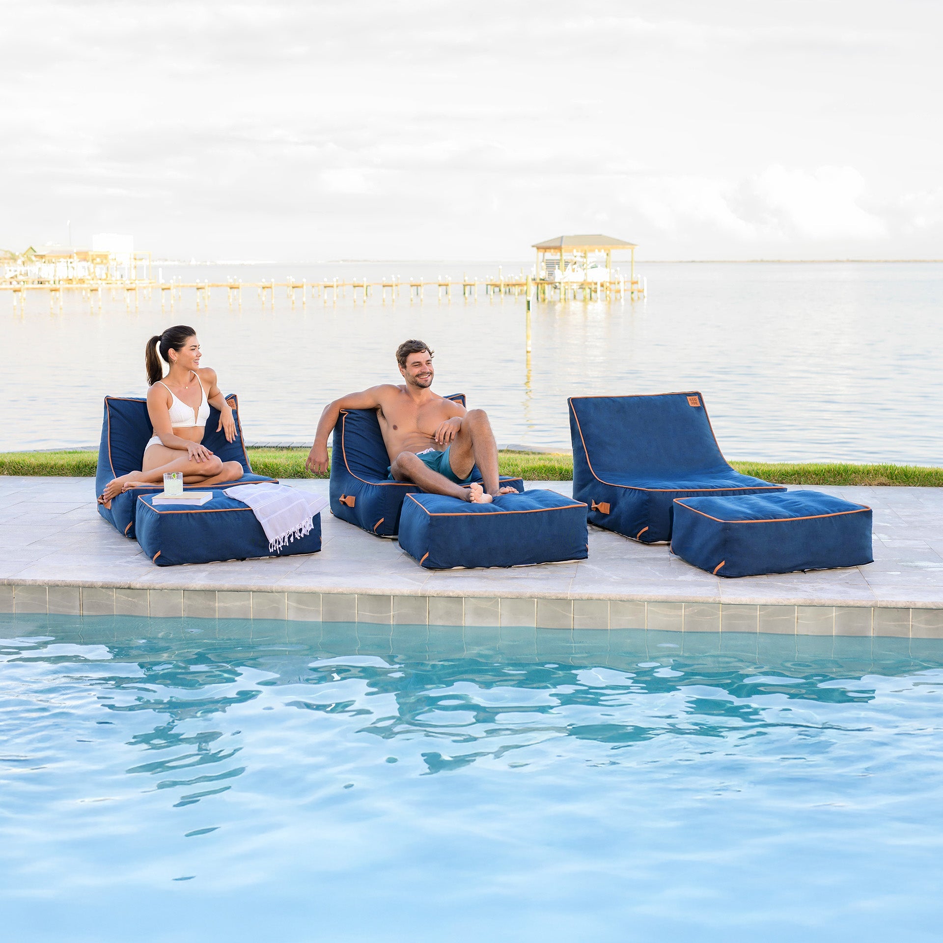 Two people sitting on blue lounge chairs by a poolside with decorative lights in the background.