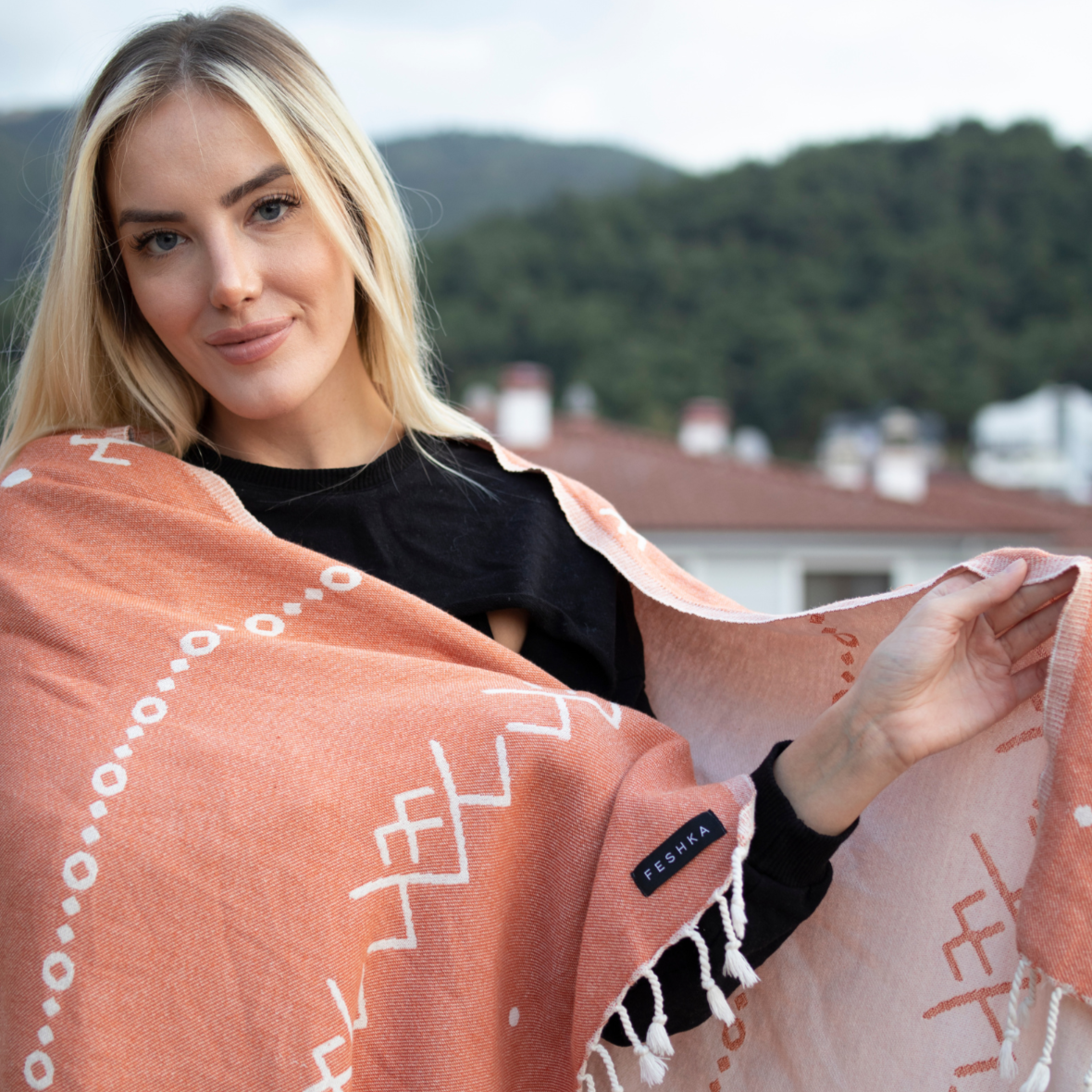 Woman holding a pink patterned towel with 'Penha' branding on a rooftop with greenery in the background.