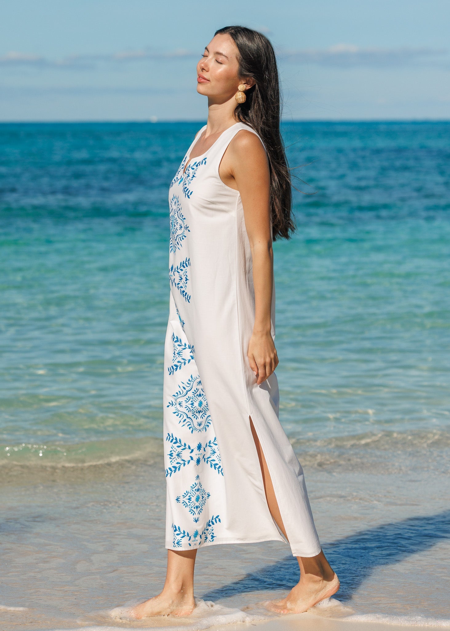 Woman in a white dress with blue patterns standing on a beach with clear blue water.
