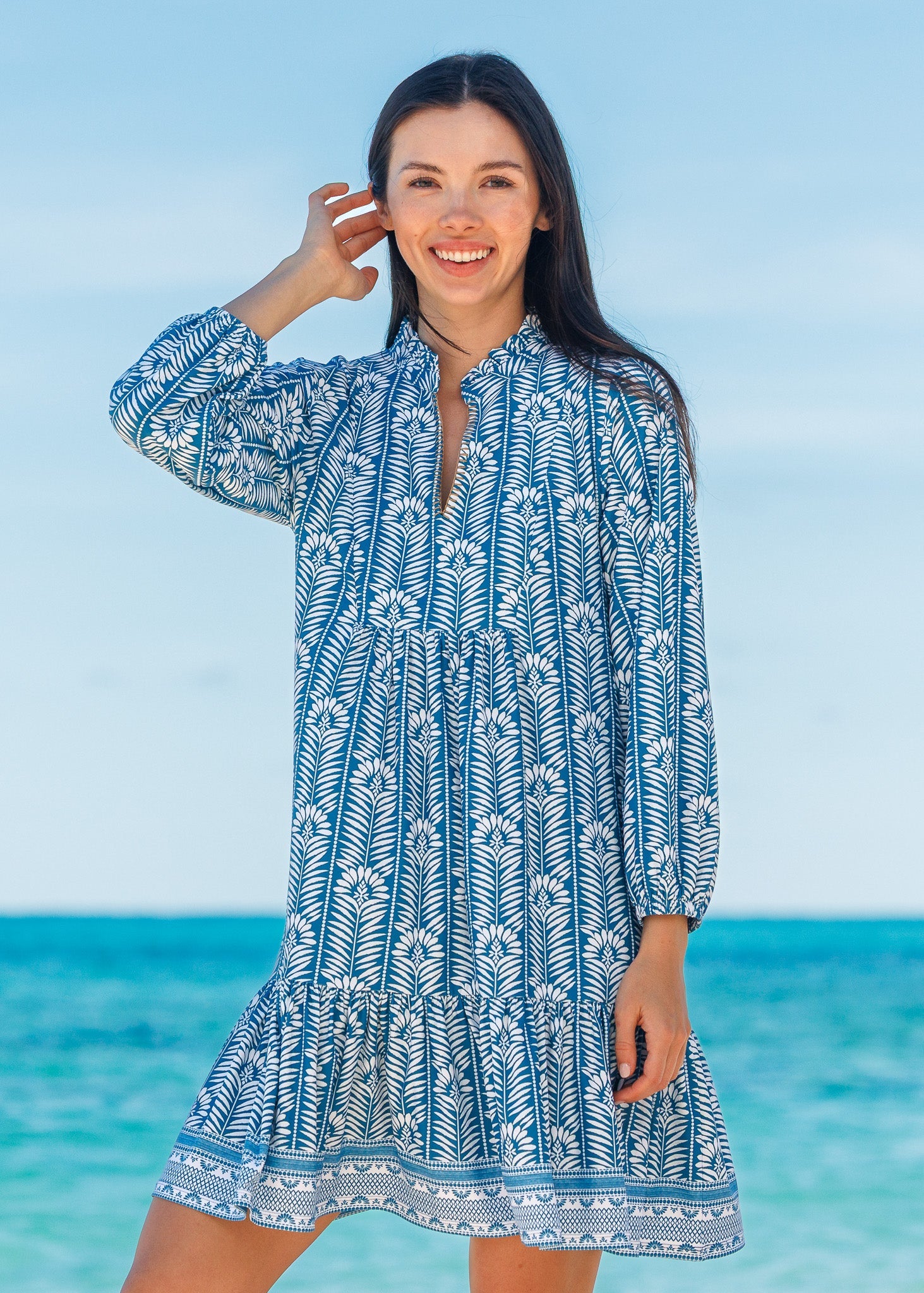 Woman wearing a blue and white patterned dress standing on a beach with clear blue water and sky.