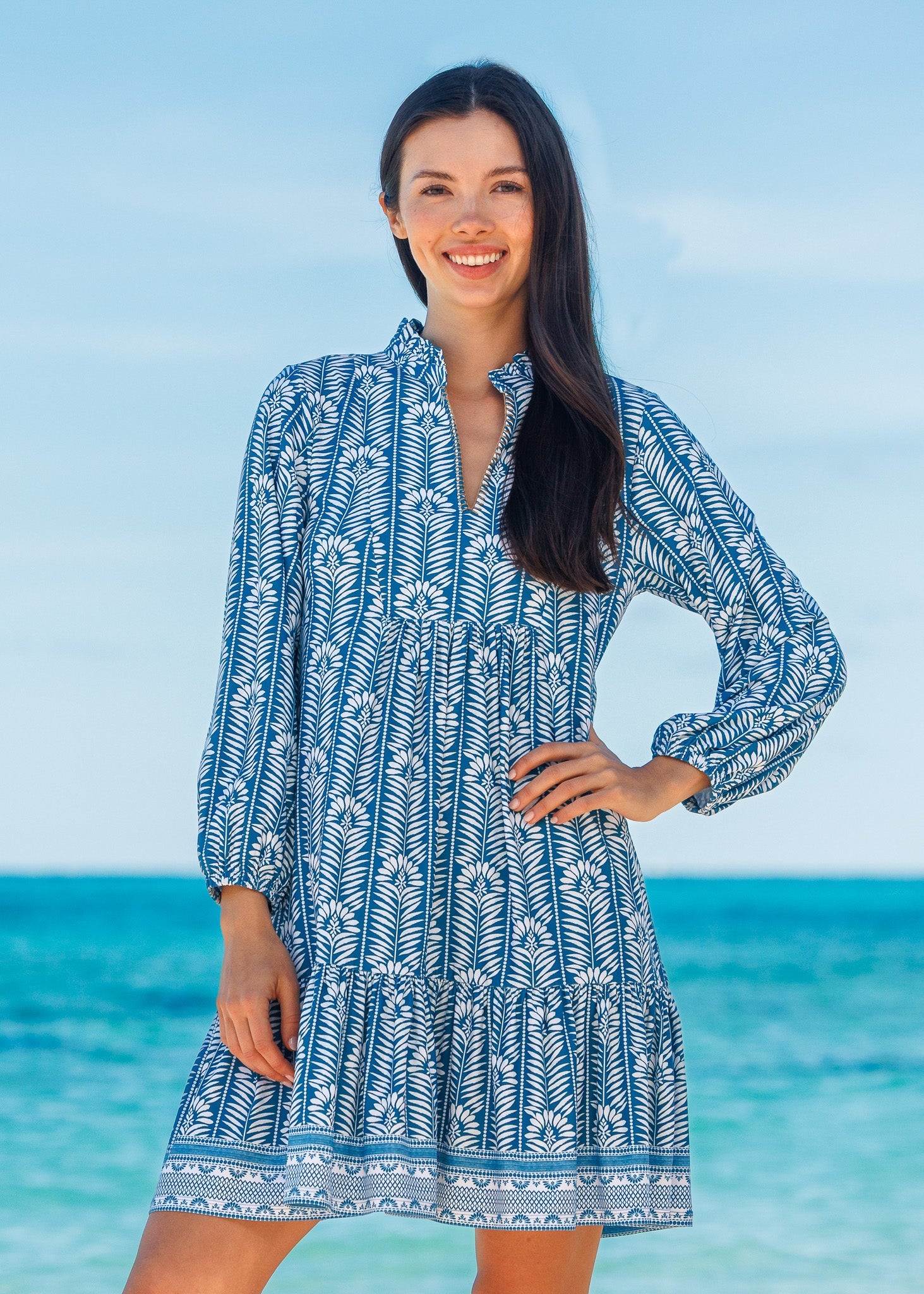 Woman wearing a blue and white patterned dress standing in front of a clear blue sky and ocean.