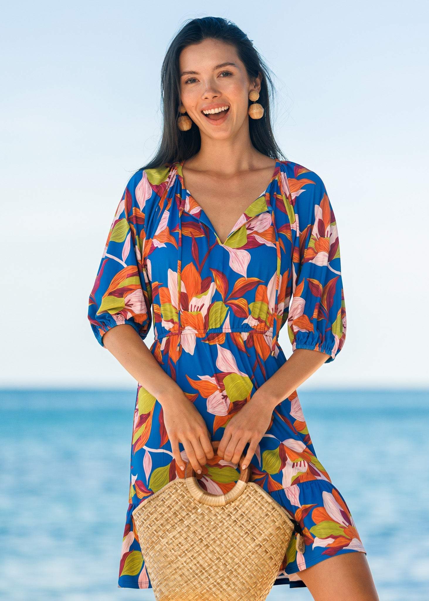 Woman wearing a colorful floral dress with a straw bag on a beach.