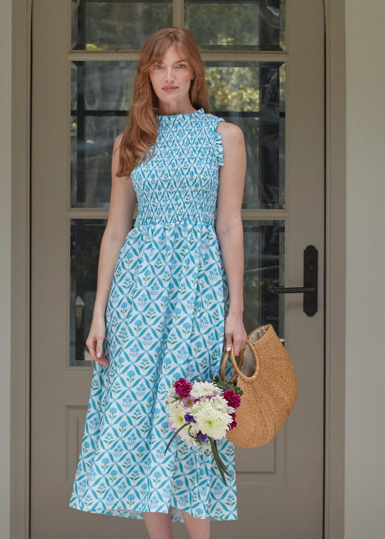 Woman in a blue patterned dress holding flowers and a straw bag in front of a glass door.