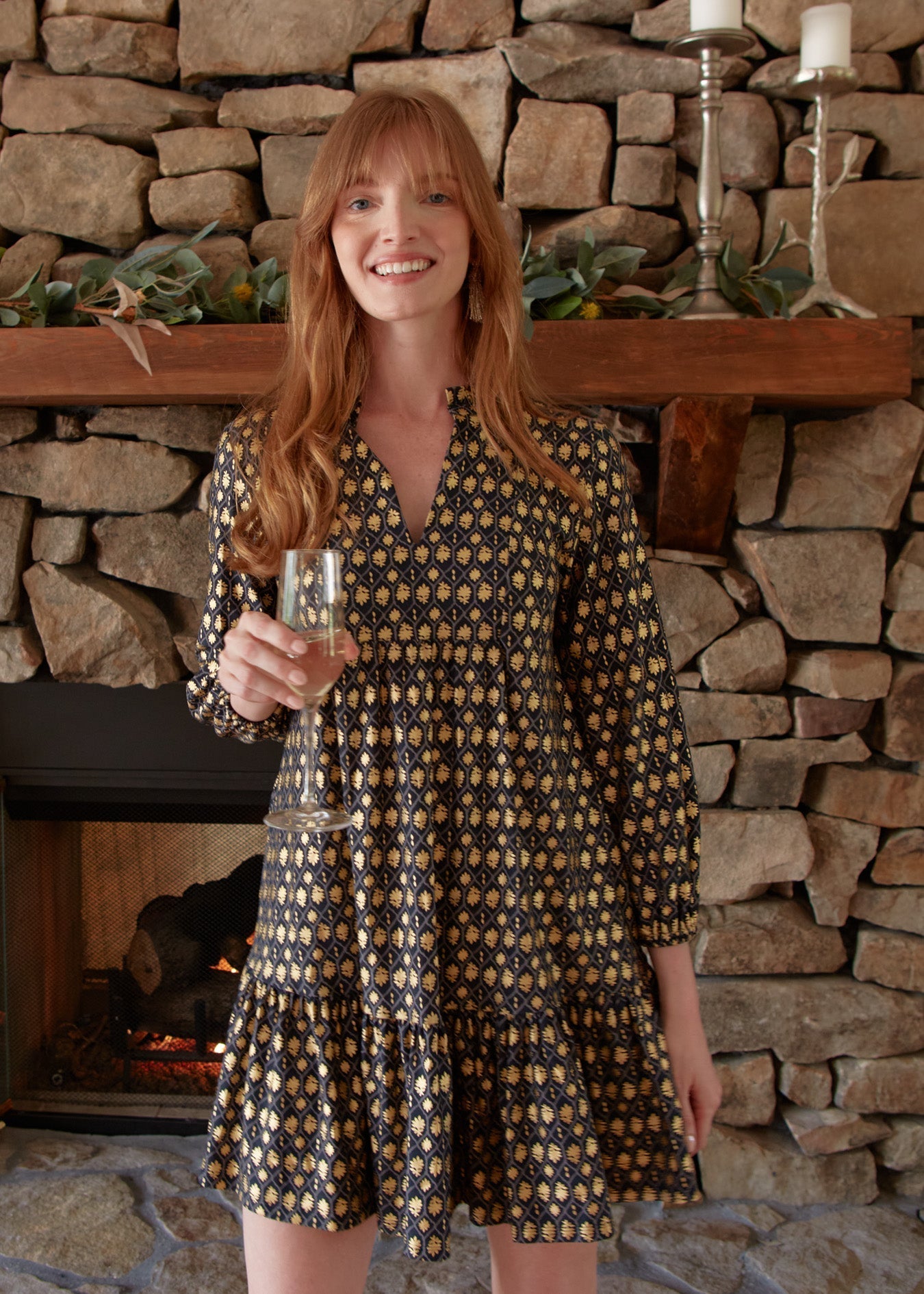 Woman in a patterned dress holding a glass in front of a stone fireplace.