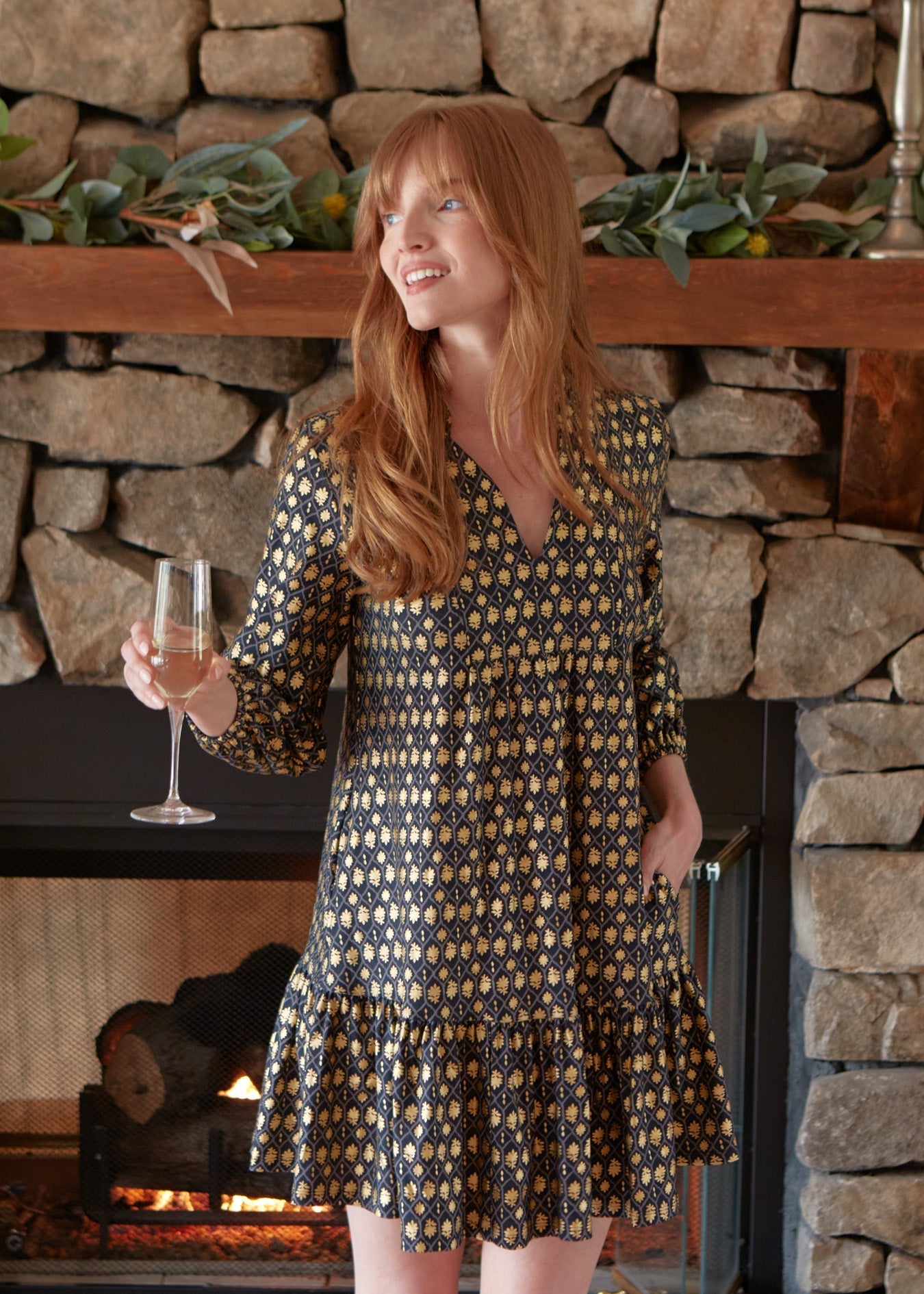 Woman in a patterned dress holding a glass of wine in front of a stone fireplace.