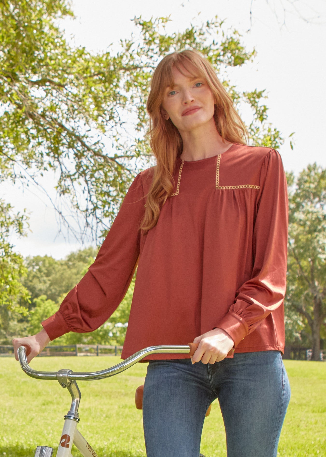 Woman in a rust-colored blouse standing next to a bicycle in a park.