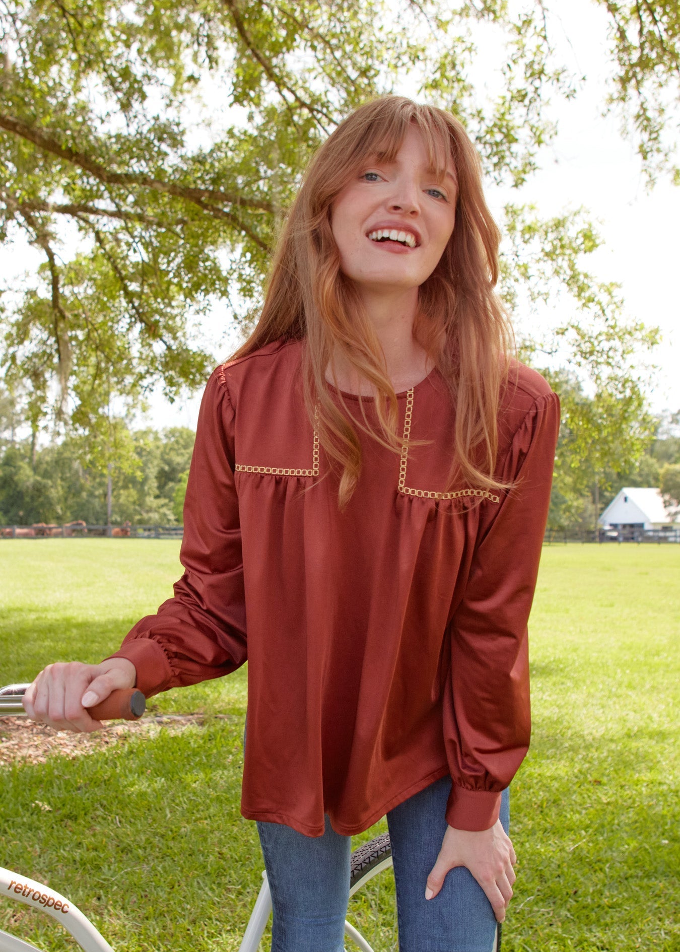 Woman in a rust-colored blouse standing outdoors with trees and grass in the background