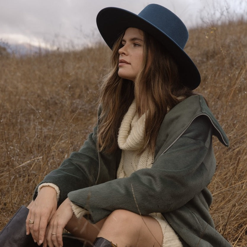 Woman sitting in a field wearing a blue hat and green coat.