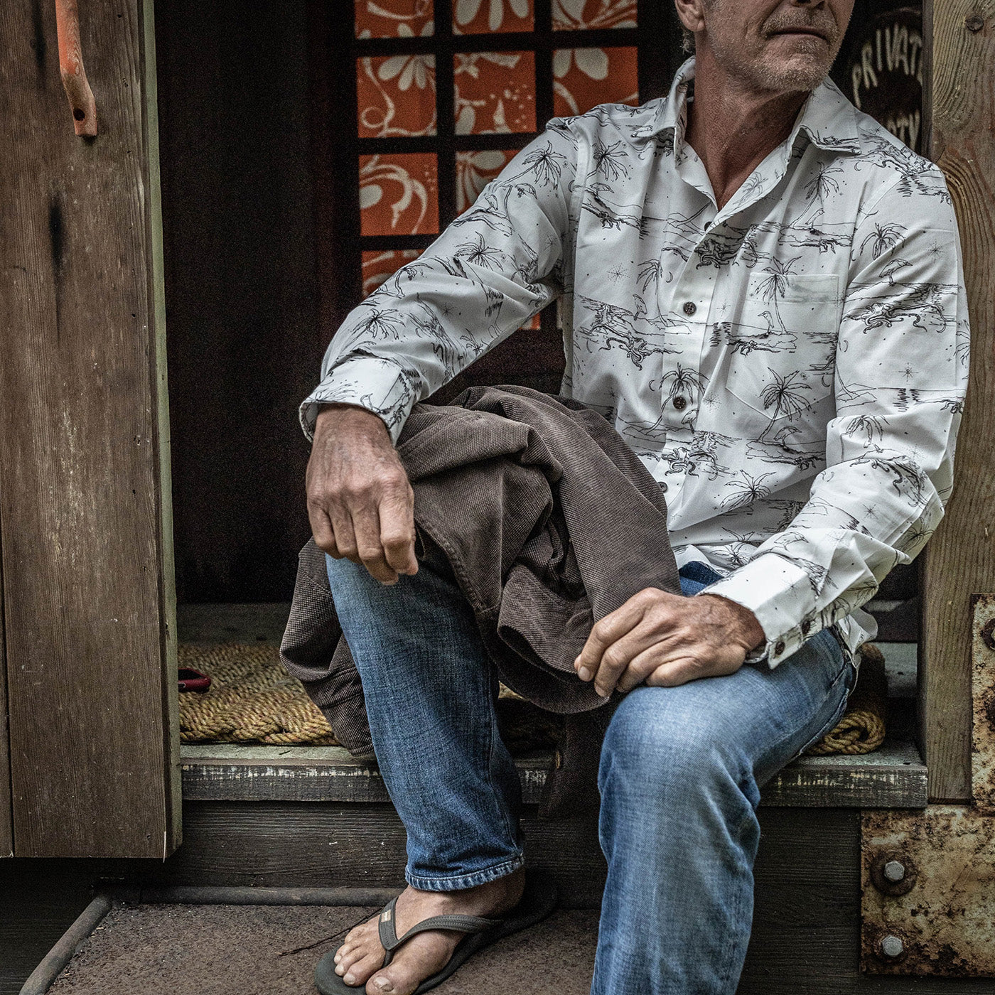 Man sitting on a step wearing a patterned shirt and jeans, with a rustic background.