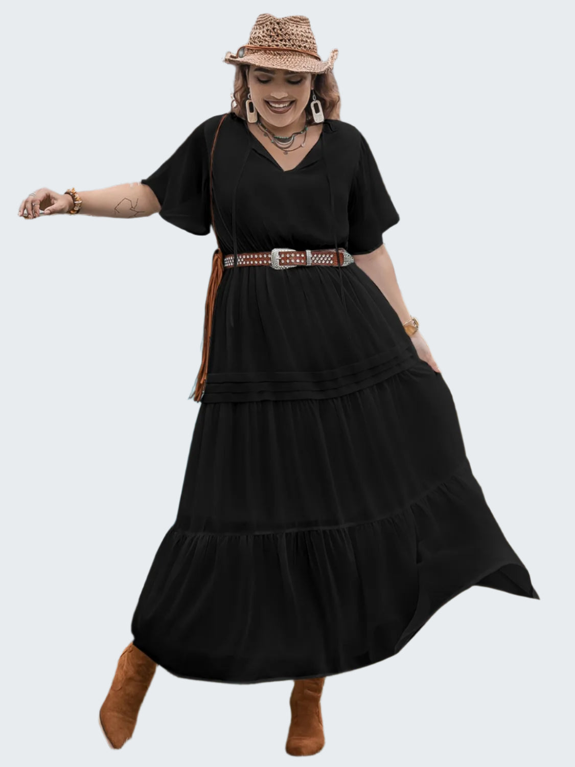 A woman standing beside a vintage van wearing a black tie neck flutter sleeve dress with a v-neckline, short sleeves, and a maxi length. She has accessorized with a brown belt, boots, and a wide-brimmed hat.