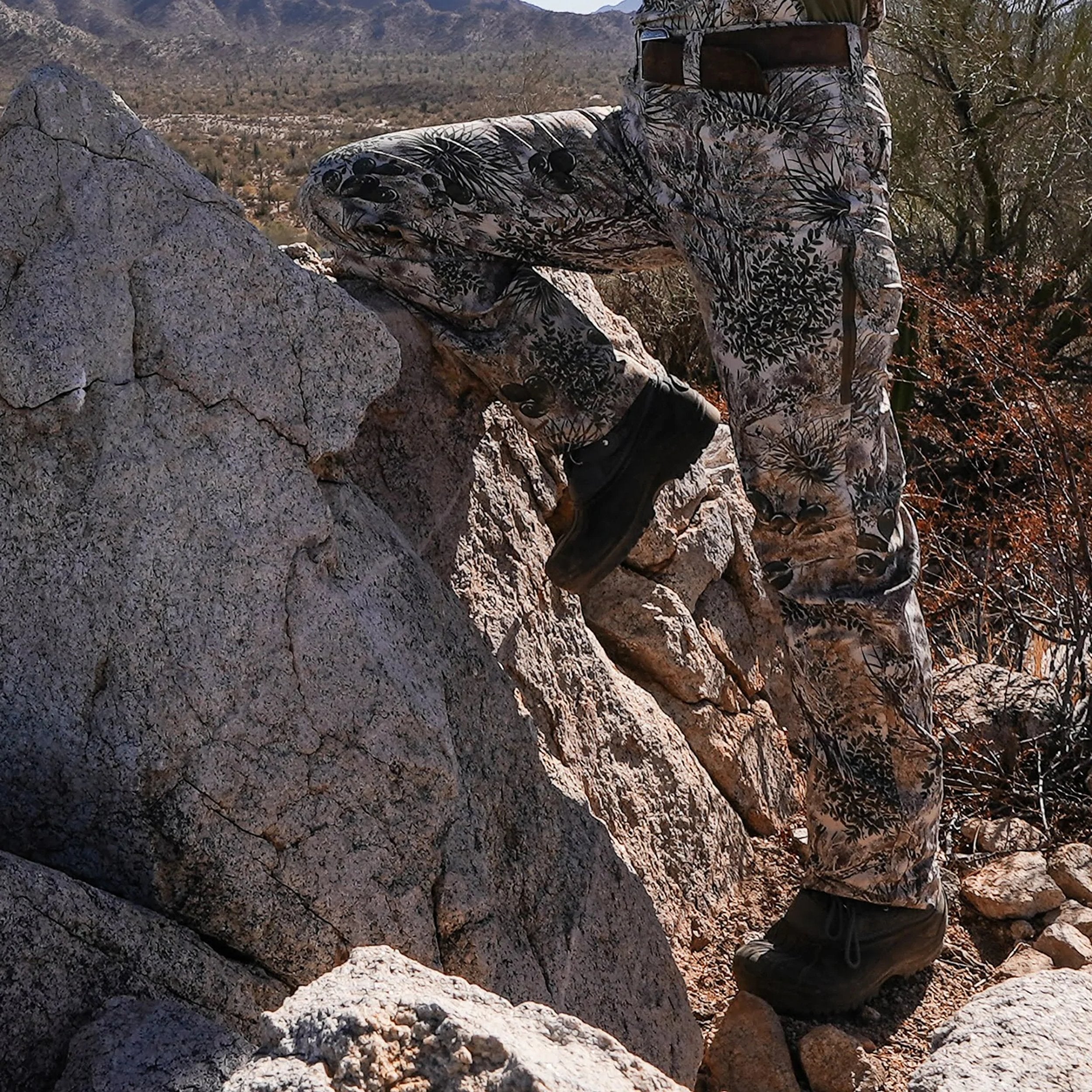 Person in camouflage attire crouching behind a rock in a desert landscape