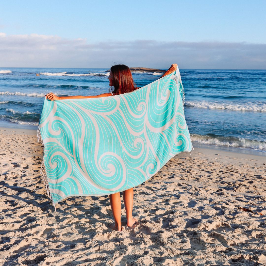 Person holding a turquoise and white patterned towel on a beach with ocean waves in the background