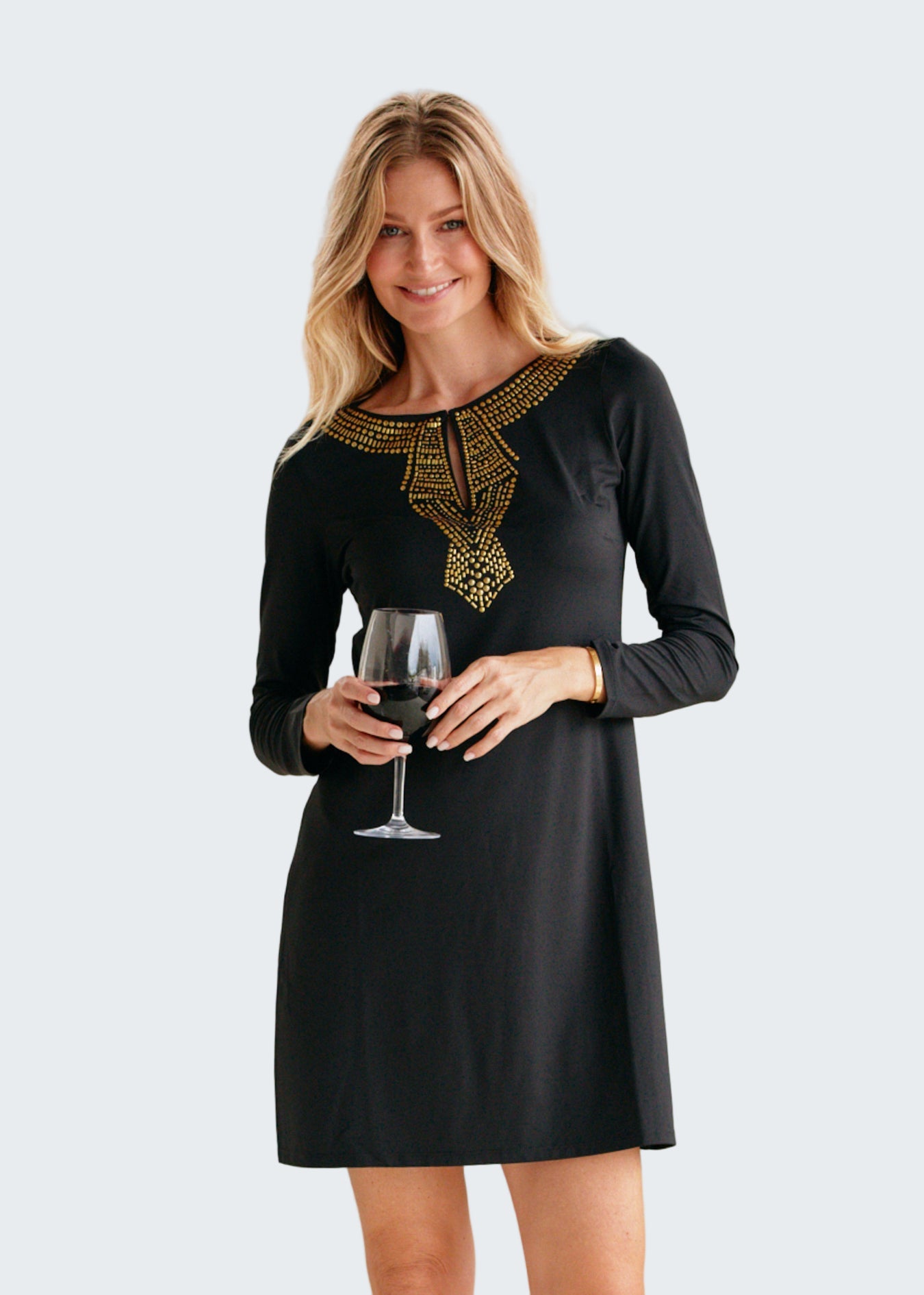 Woman in a black dress with gold embroidery holding a glass of red wine against a beige background