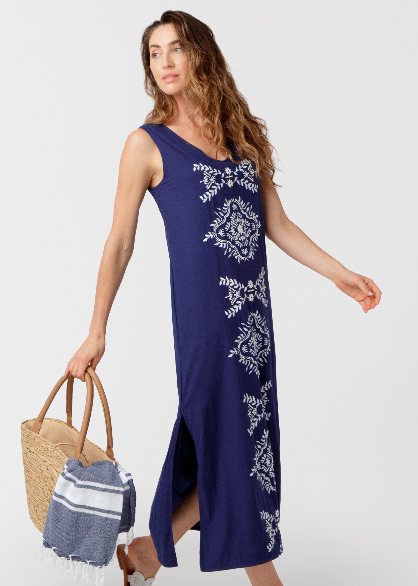 Woman wearing a navy blue dress with white patterns, holding a straw bag and a towel on a white background