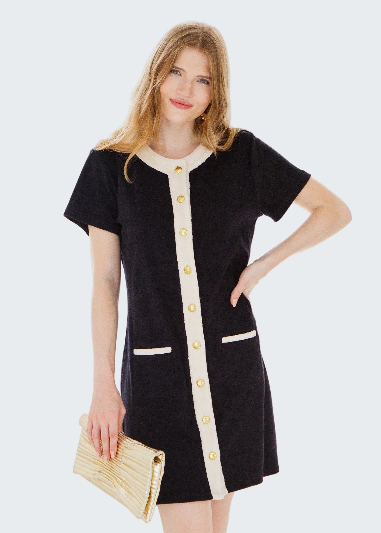 Woman wearing a black dress with white buttons and trim, holding a beige clutch in front of a window.